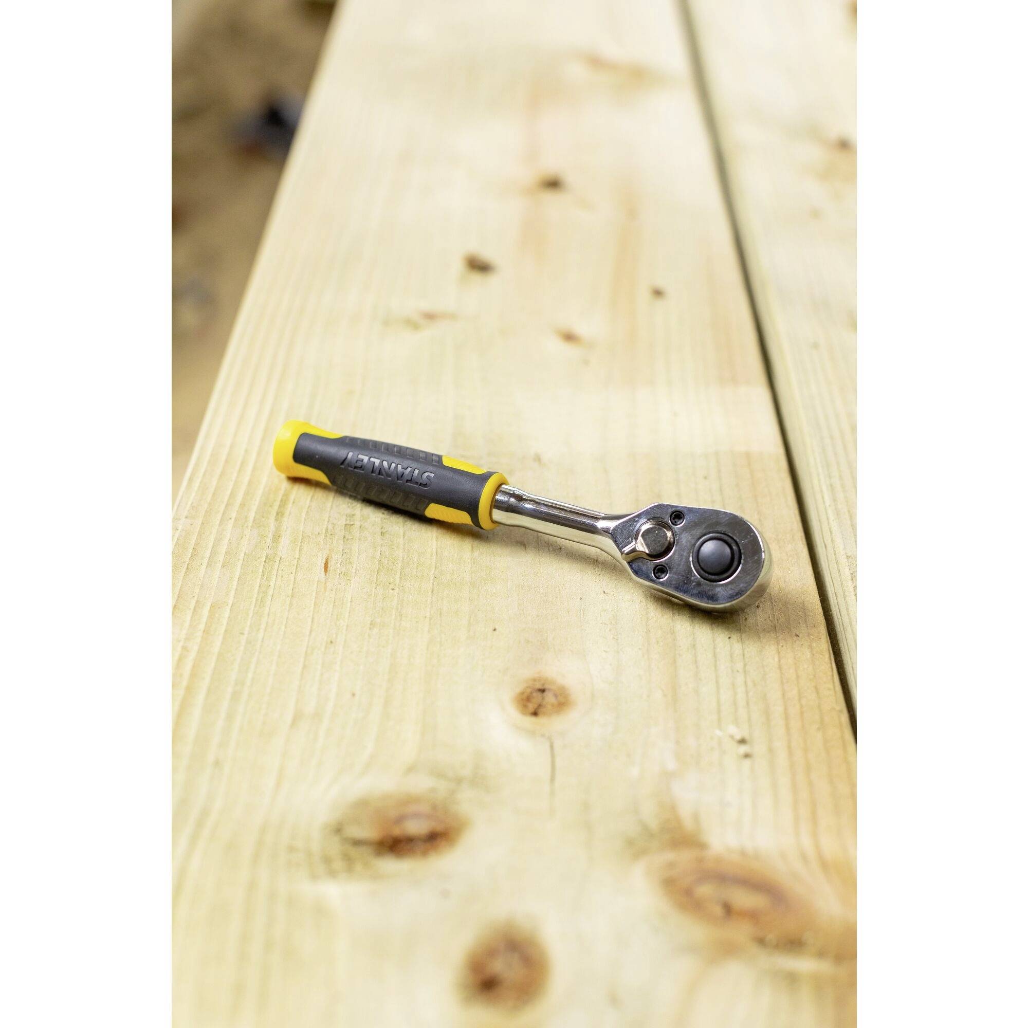 Klucz z mechanizmem zapadkowym z żółto-czarnym uchwytem leży na drewnianym belkowaniu. Belka drewniana wykazuje lekkie ślady zużycia.