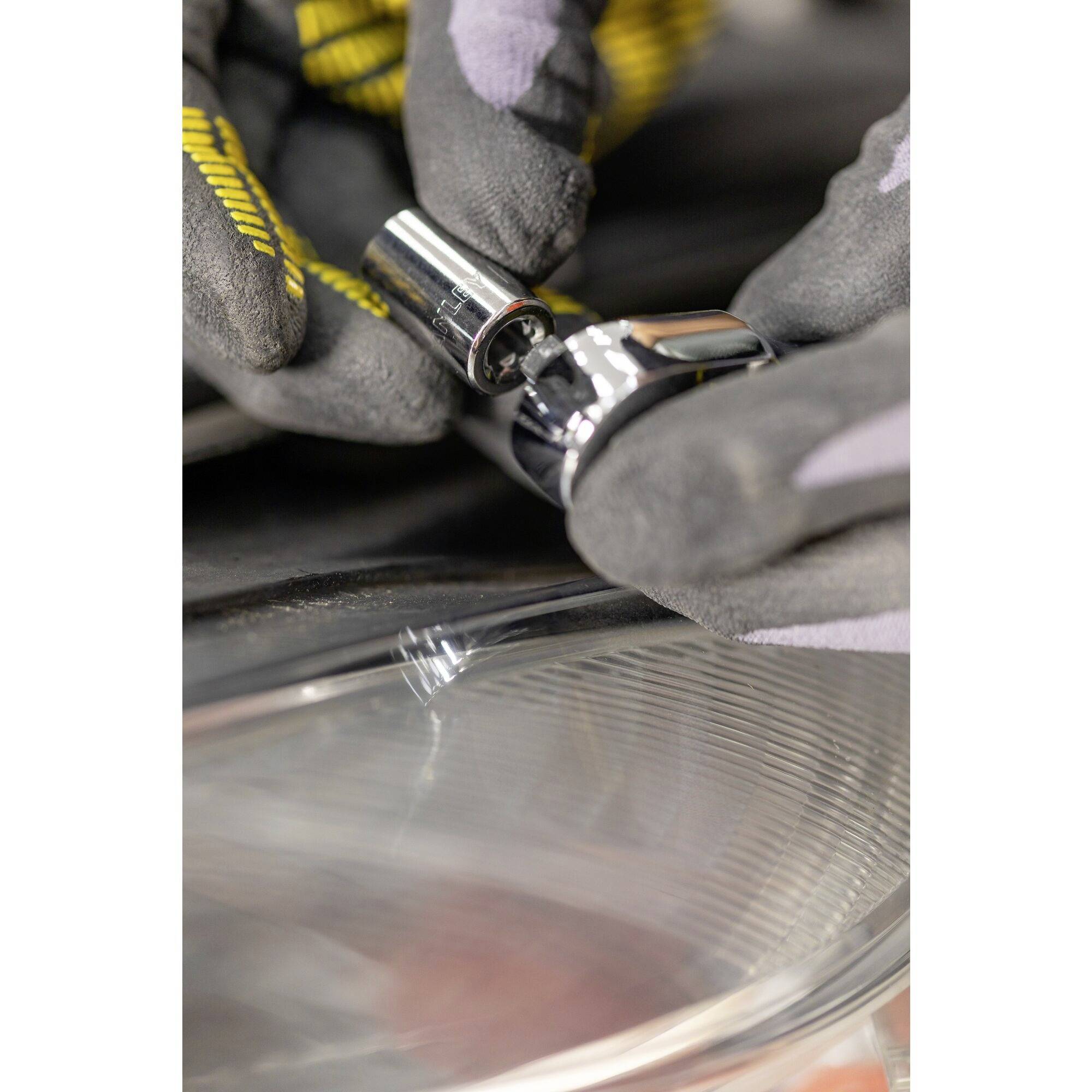 Osoba w rękawiczkach naprawia lub montuje część samochodową za pomocą narzędzia śrubowego. Skupiona jest na pracy przy reflektorze.