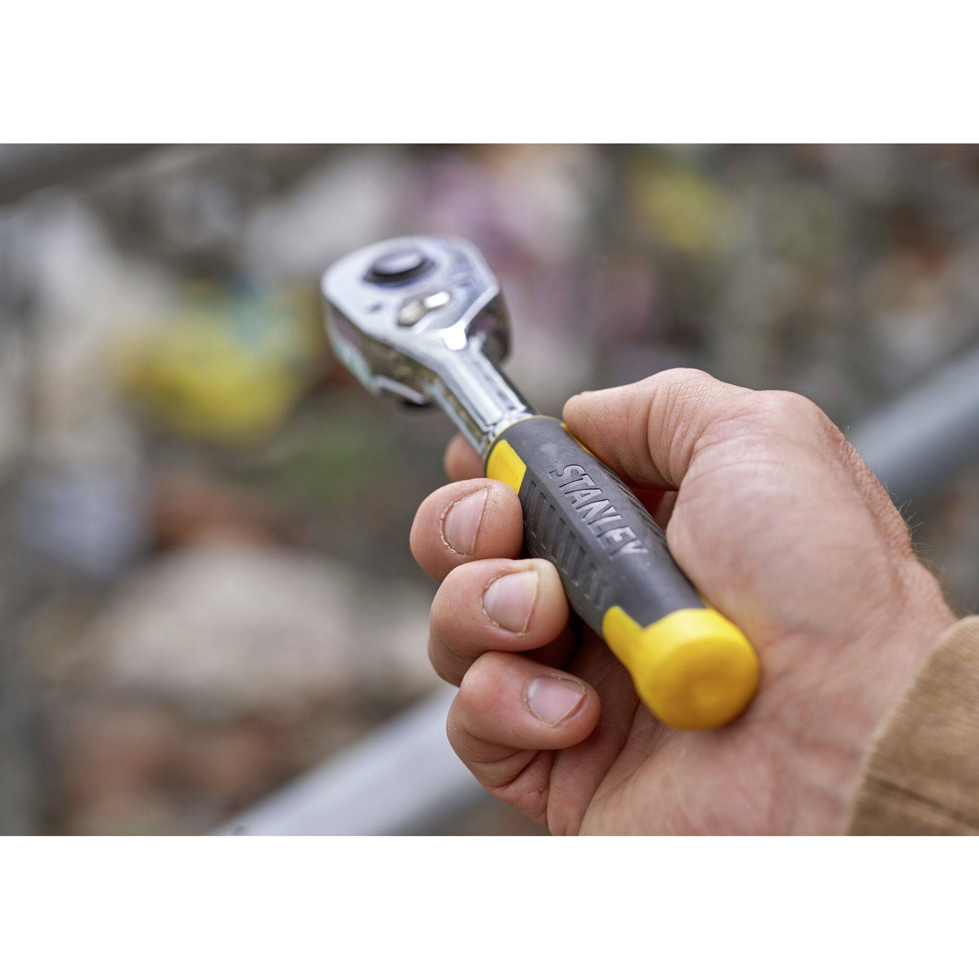 Dłoń trzyma klucz do grzechotki z żółtą rękojeścią, w tle rozmyte tło. Główny fokus na narzędziu do prac ręcznych.