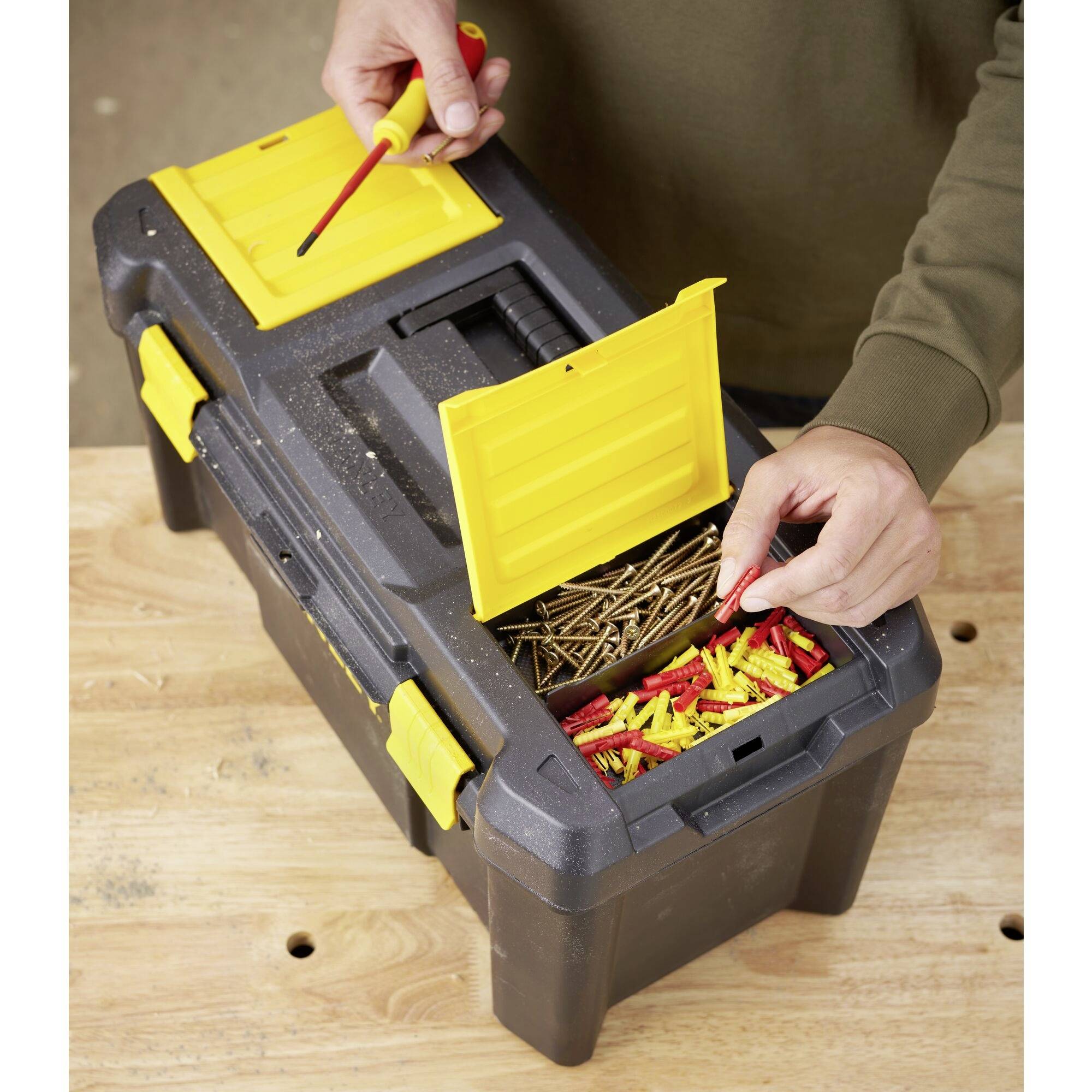 Osoba otwiera skrzynkę narzędziową z żółtą szufladą, wypełnioną śrubami i kołkami, na drewnianym blacie roboczym.