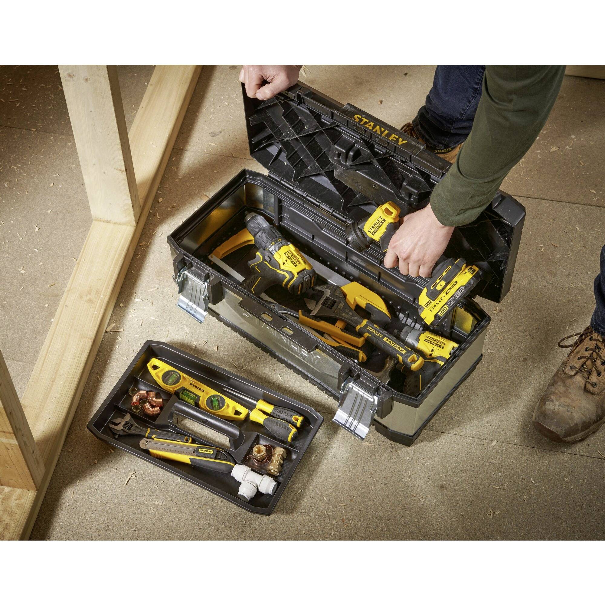 Otwarty skrzynka narzędziowa na drewnianej podłodze, wypełniona różnymi narzędziami, w tym wiertarką i poziomicą. Dwie dłonie manipulujące lub rozmieszczające narzędzia.