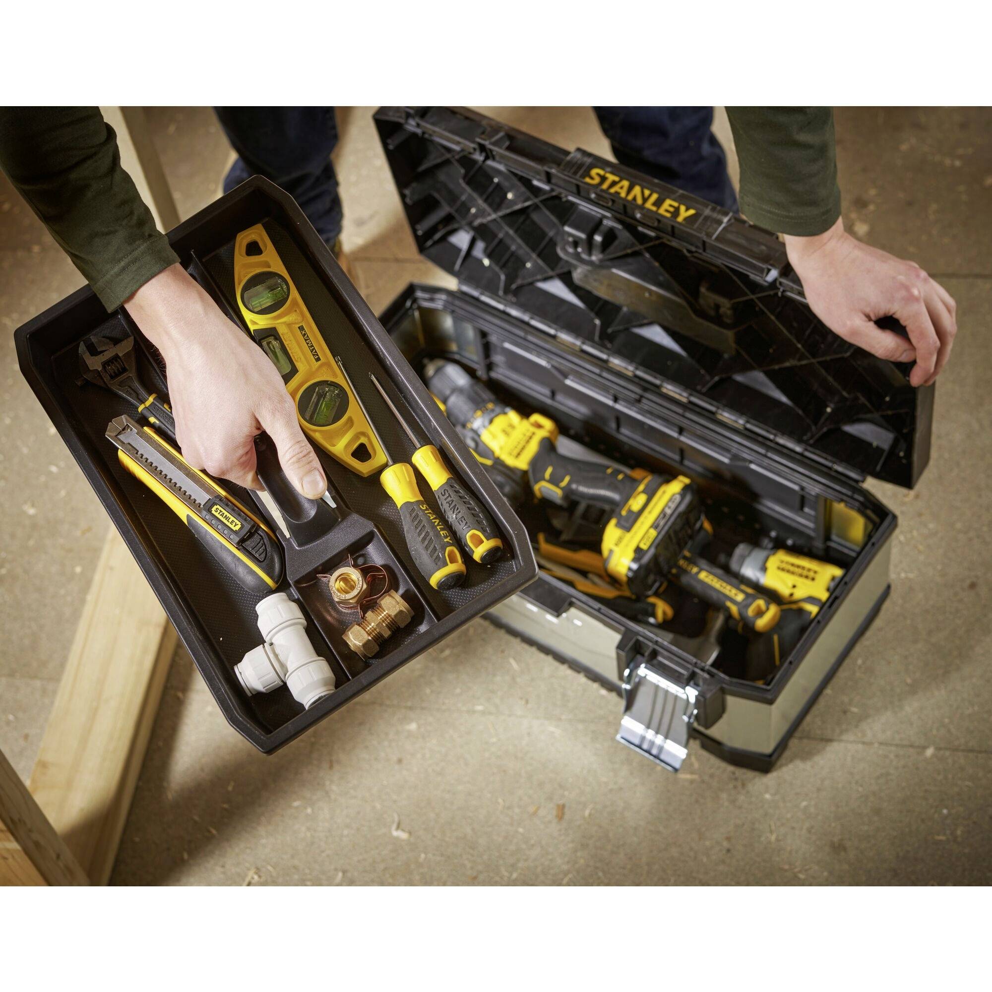 Dwa otwarte walizki z narzędziami zawierające różne przyrządy takie jak wiertarka, poziomnica i śrubokręt na drewnianej podłodze. Ręce trzymają jedną z szuflad.