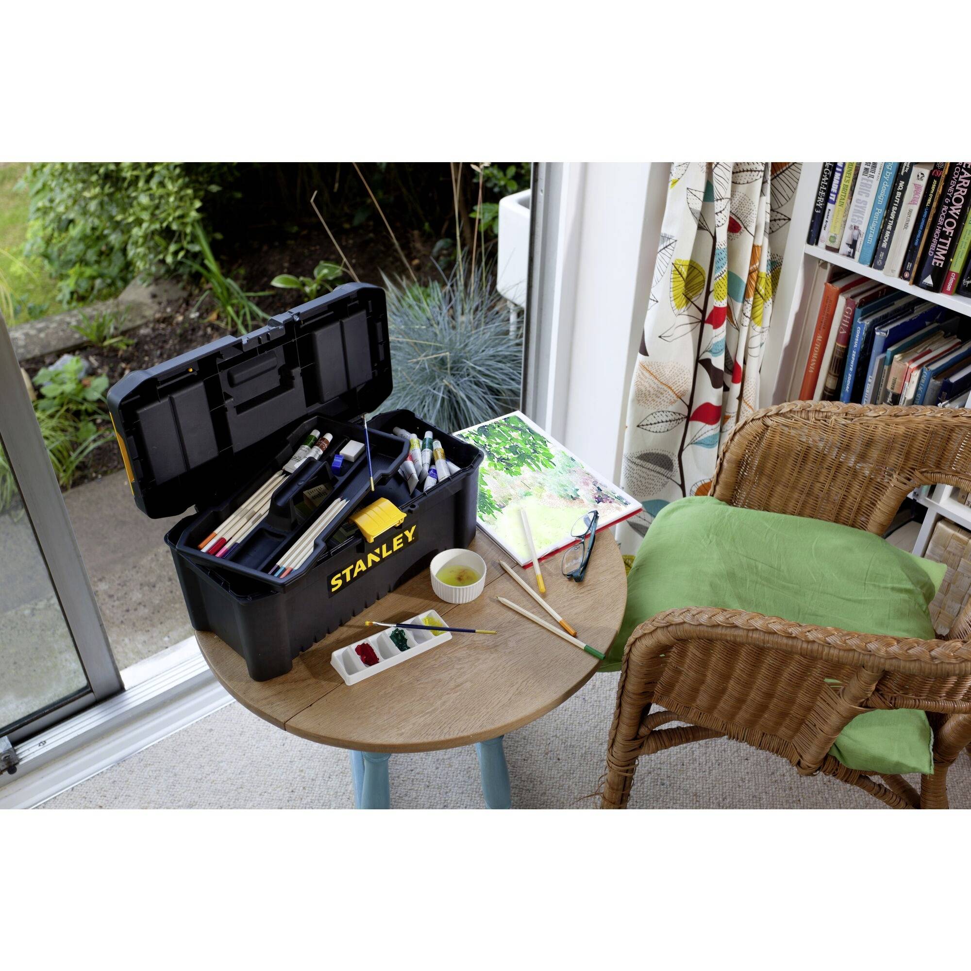 Otwarty przybornik z przyborami plastycznymi stoi na stole obok zielonego krzesła i książki do kolorowania, w jasnym pomieszczeniu.