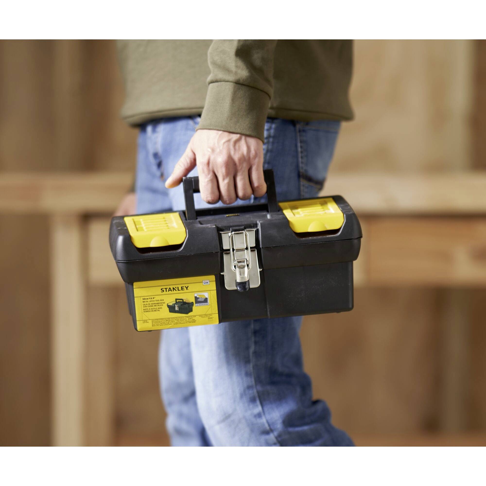 Osoba niesie czarną skrzynkę narzędziową z żółtymi zamknięciami w ręku, a w tle widać plac budowy.