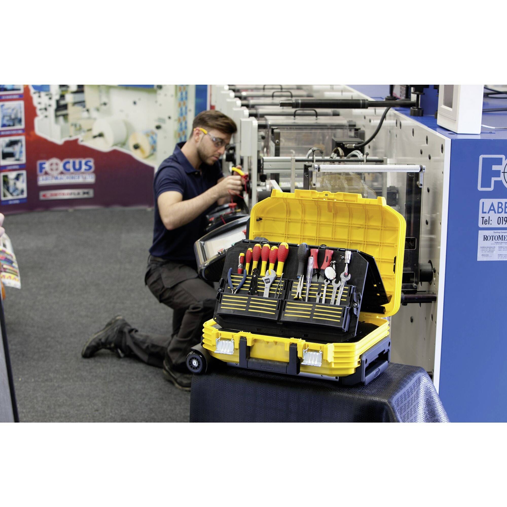 Technik pracuje przy maszynie. Na pierwszym planie stoi otwarty żółty kufer narzędziowy z różnymi narzędziami.