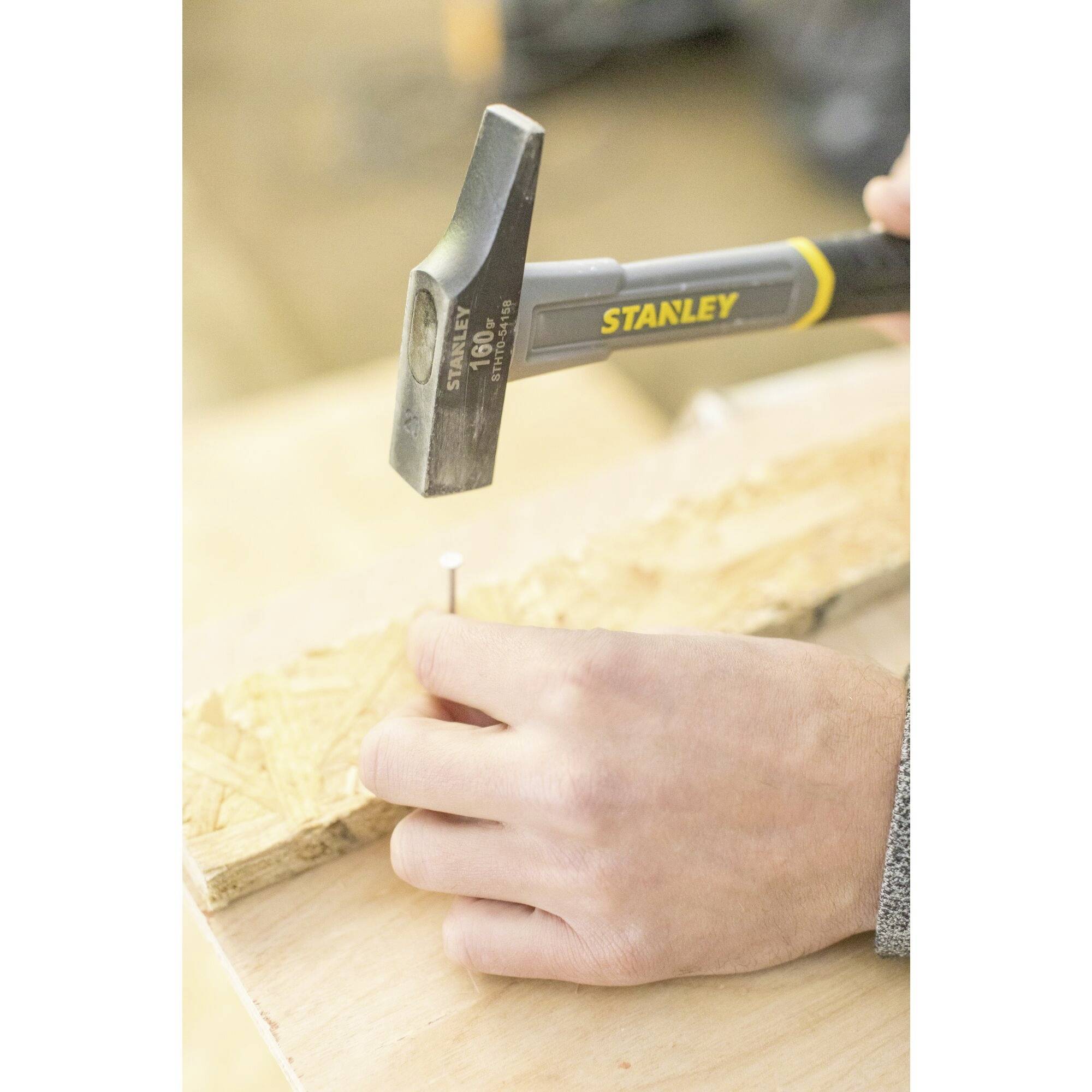 Osoba wbija gwoździa w kawałek drewna. Używany jest młotek marki Stanley. Ręka trzyma gwoździa stabilnie.