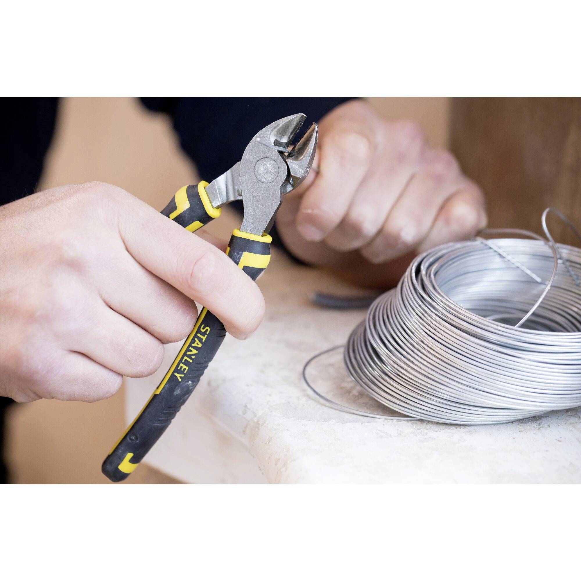 Osoba tnie drut żółtymi szczypcami. Obok leży rolka drutu na stole.