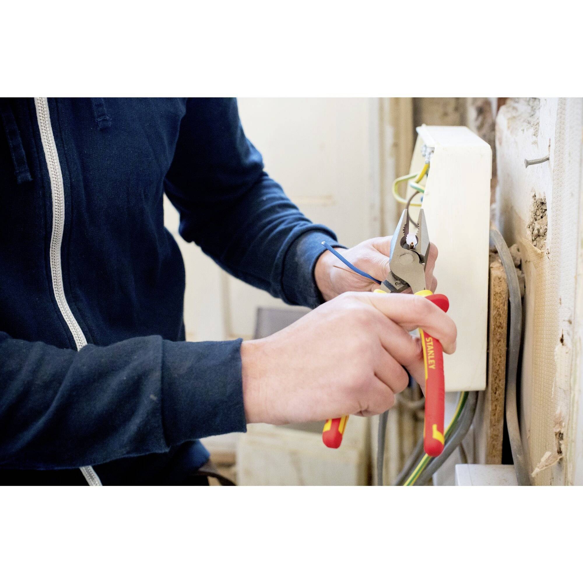 Osoba instaluje lub naprawia przewody elektryczne w ścianie. Używa szczypiec i ma na sobie ciemną bluzę.