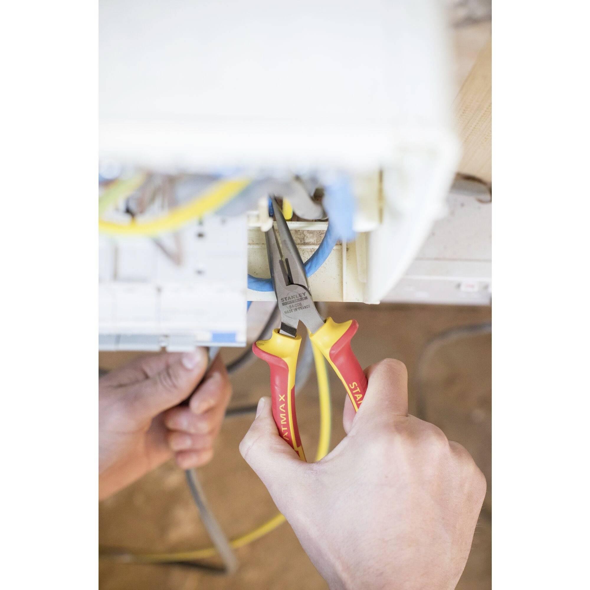 Osoba pracuje szczypcami izolacyjnymi przy przewodach elektrycznych w skrzynce rozdzielczej. Szczypce mają czerwone i żółte uchwyty.