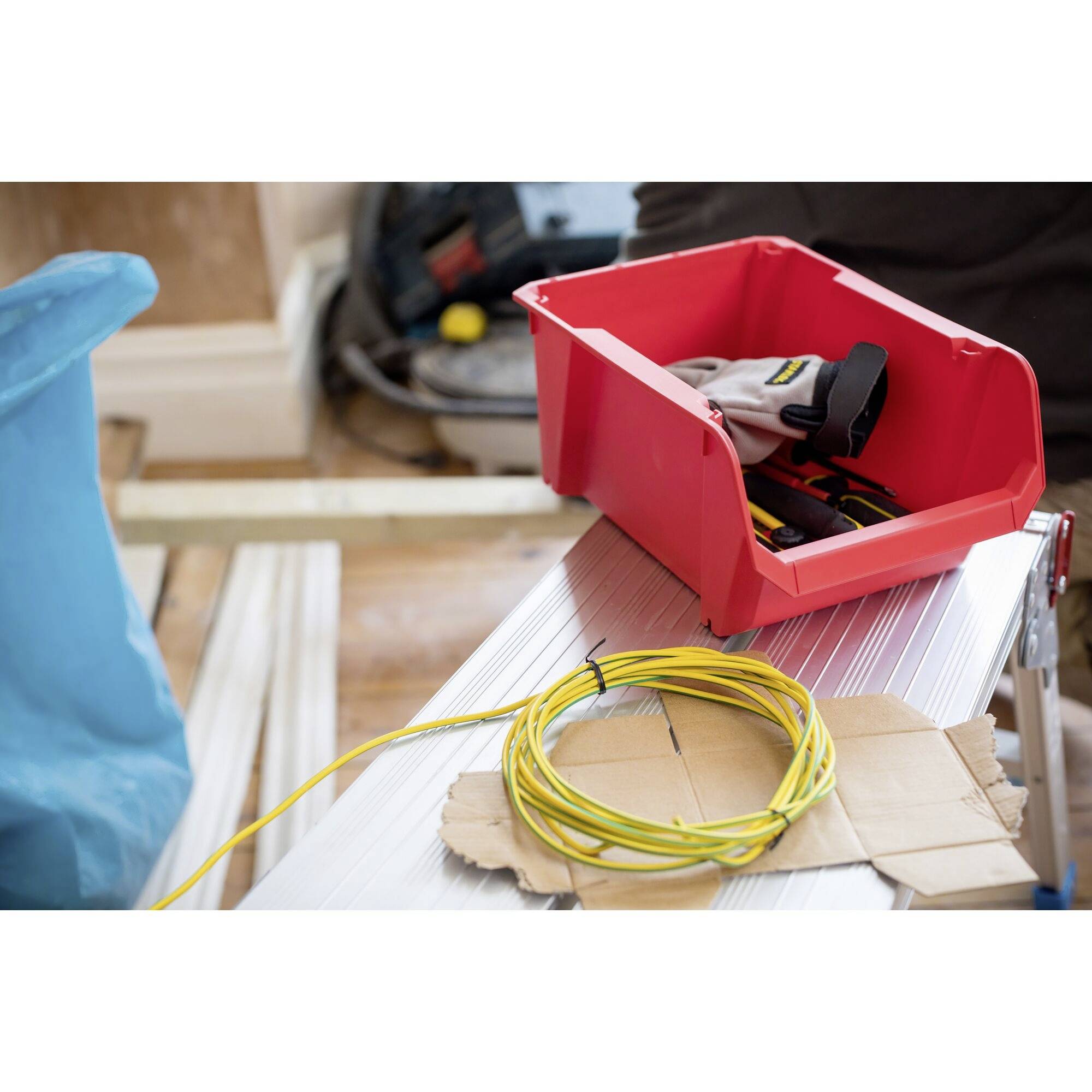 Czerwona skrzynka narzędziowa z rękawicami roboczymi i kablami leży na stole. W tle widoczne są kolejne narzędzia.
