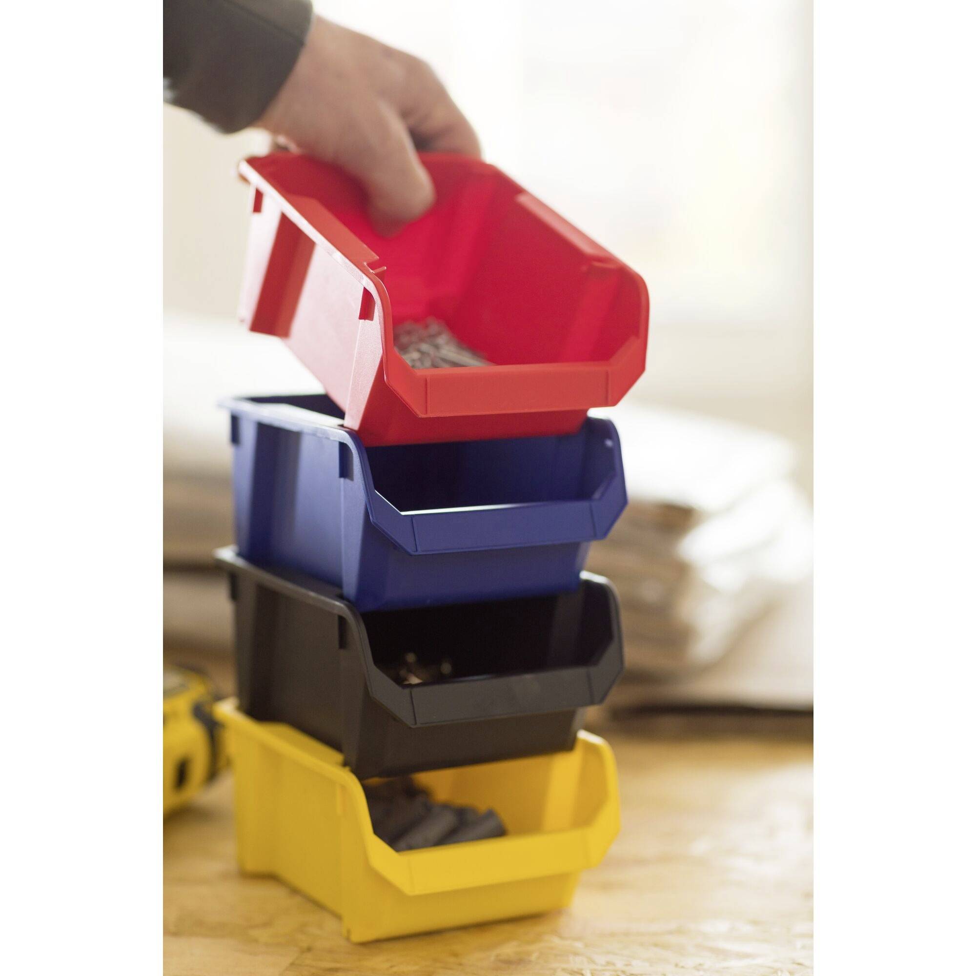 Jedna ręka trzyma czerwone pudełko magazynowe nad trzema ułożonymi jeden na drugim pudełkami w kolorach niebieskim, czarnym i żółtym. Zawierają one luźne śruby.