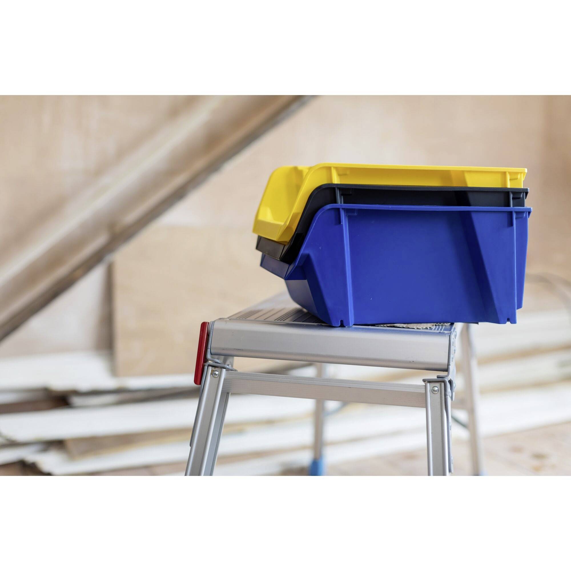 Niebieskie i żółte pudła do przechowywania są ułożone na aluminiowej drabinie w odremontowanym pomieszczeniu. W tle widać niedokończone ściany.