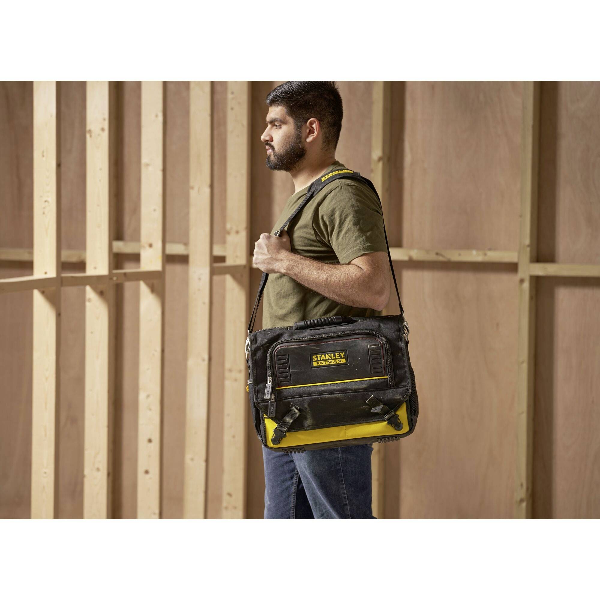 Mężczyzna nosi czarno-żółtą torbę na narzędzia marki 'STANLEY' przewieszoną przez ramię w niedokończonym pomieszczeniu.