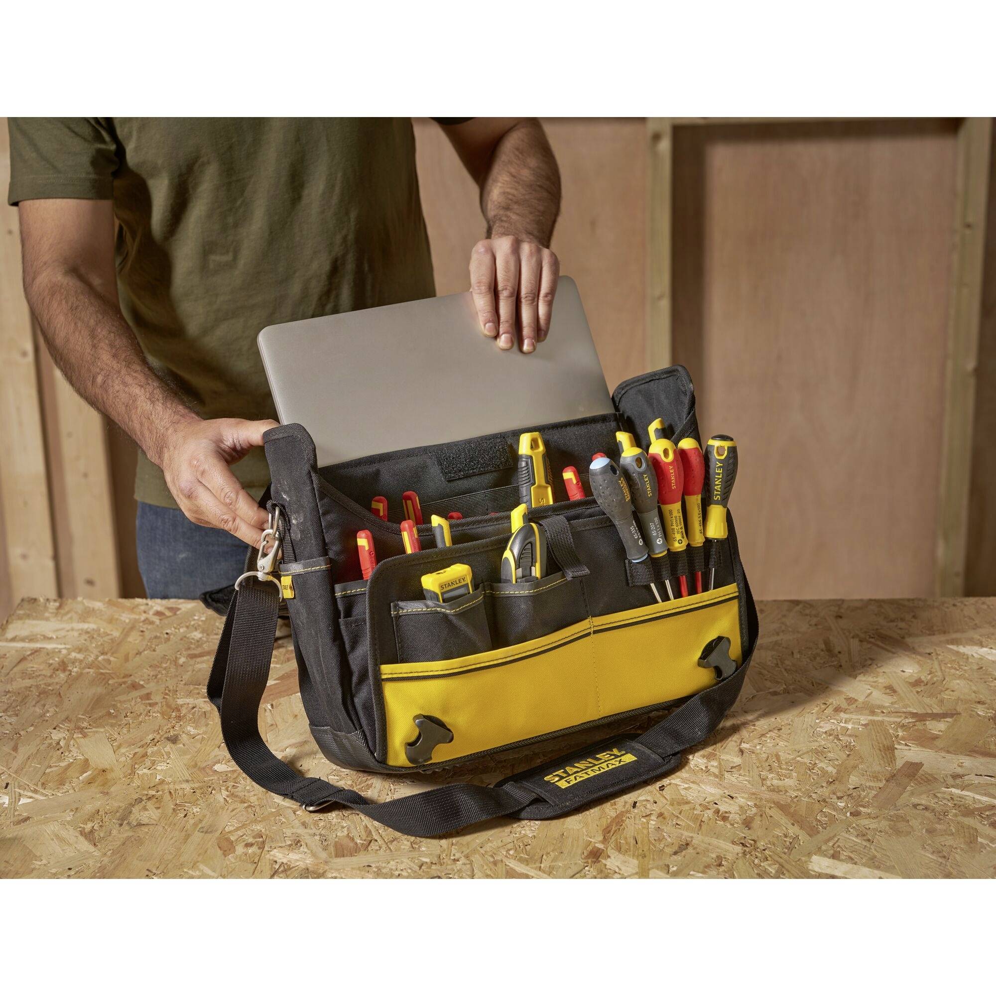Osoba wkłada laptopa do czarnej torby narzędziowej z żółtymi akcentami. Torba zawiera śrubokręty i szczypce.