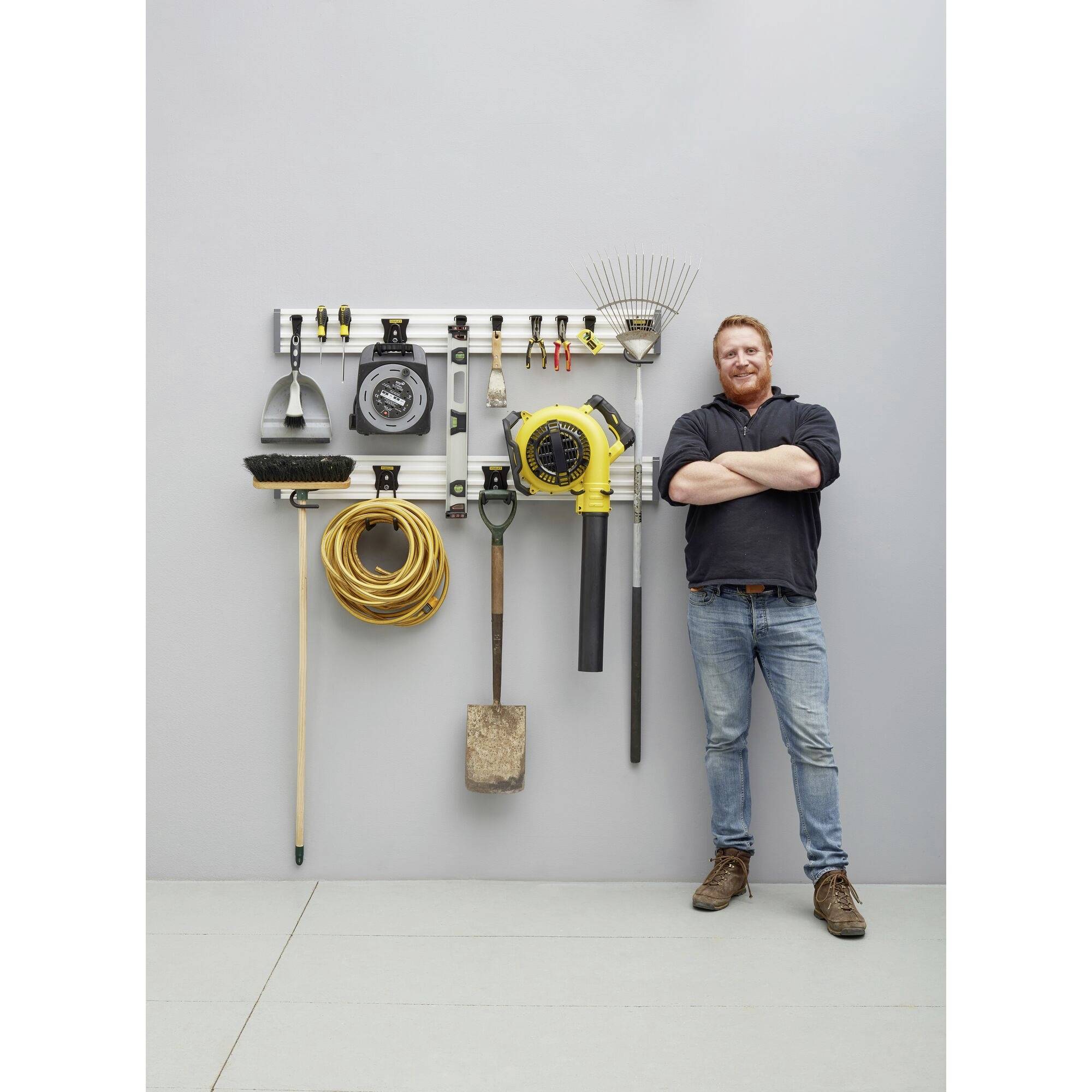 Mężczyzna stoi uśmiechnięty obok ściany, na której wiszą narzędzia ogrodowe, w tym grabie, wąż ogrodowy i łopata.