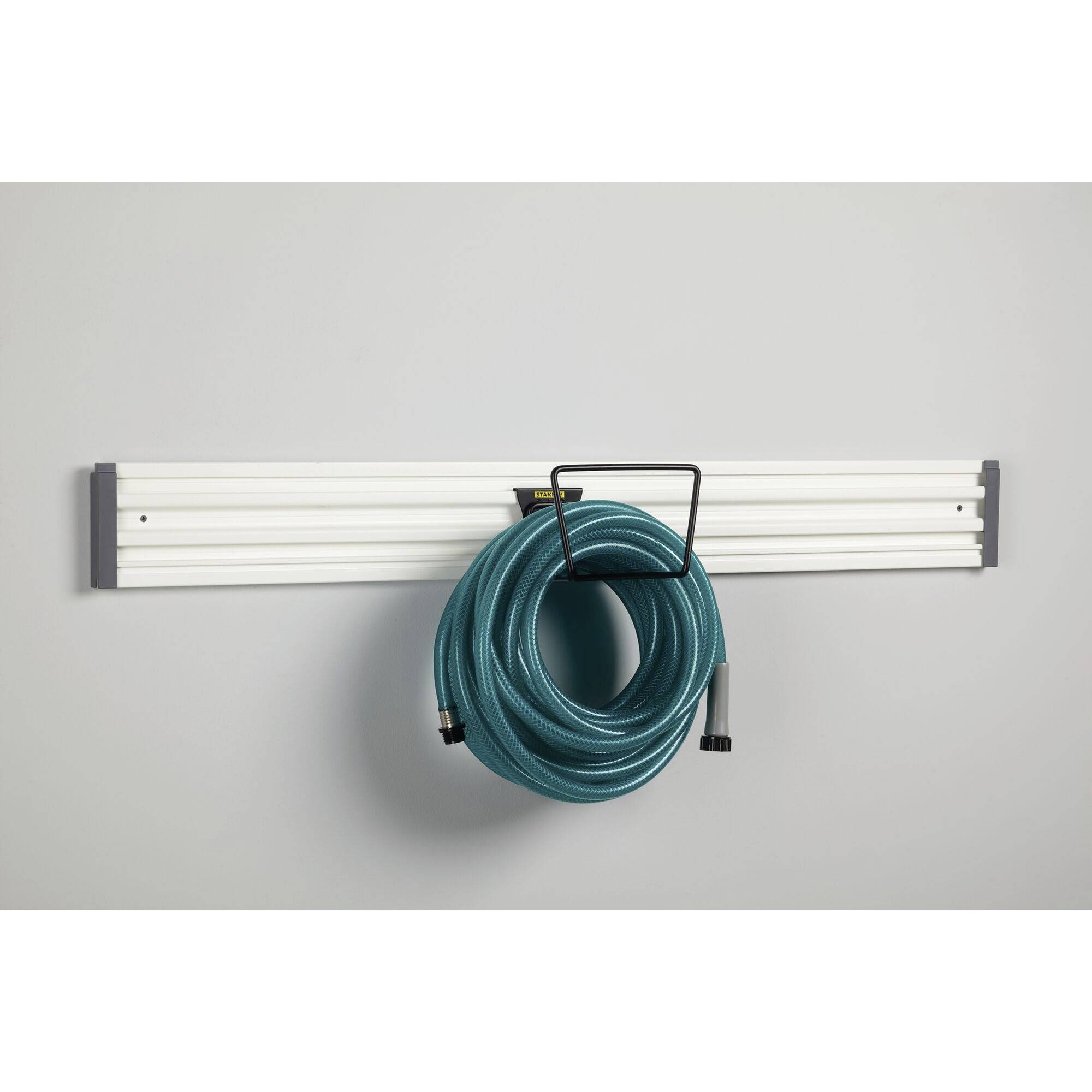 Zielony wąż ogrodowy jest starannie zawieszony na białym uchwycie ściennym przeznaczonym do przechowywania akcesoriów ogrodowych.