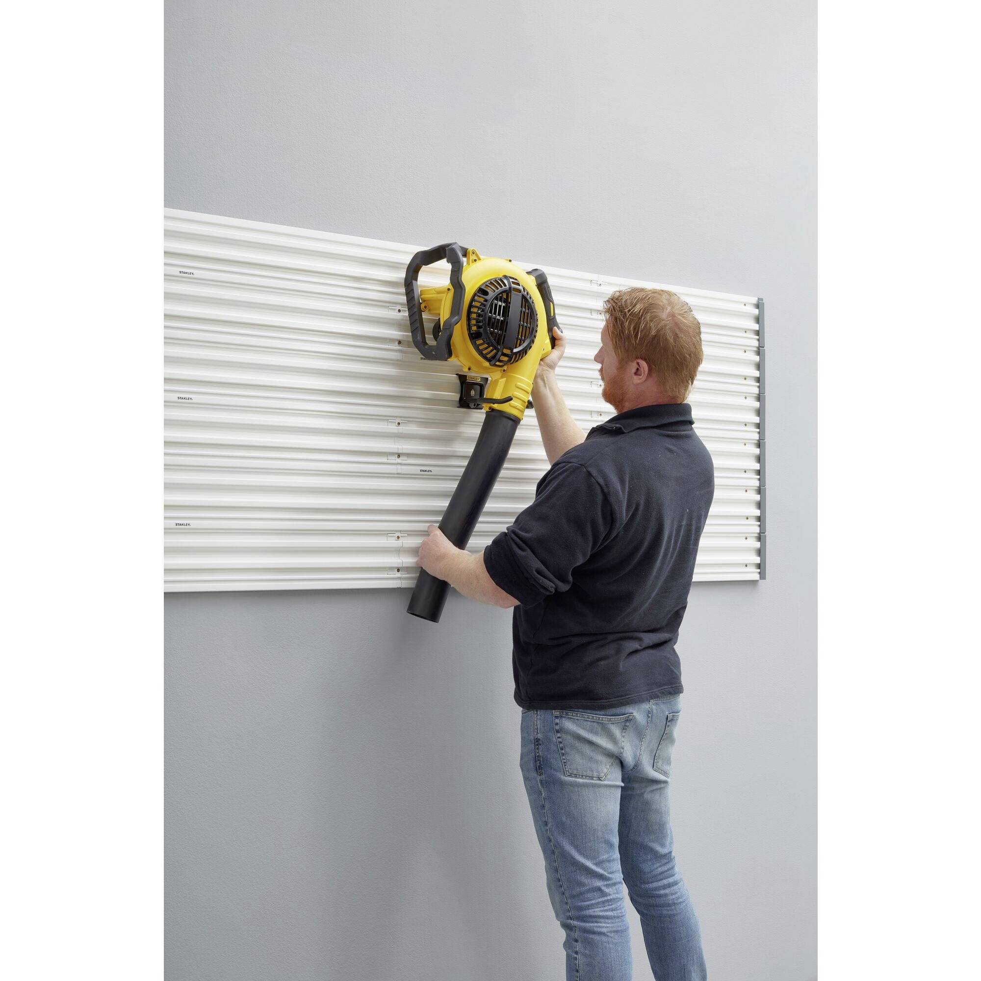 Osoba stoi przed ścianą i trzyma żółte urządzenie z czarną rurą, przypuszczalnie dmuchawę lub podobne narzędzie.
