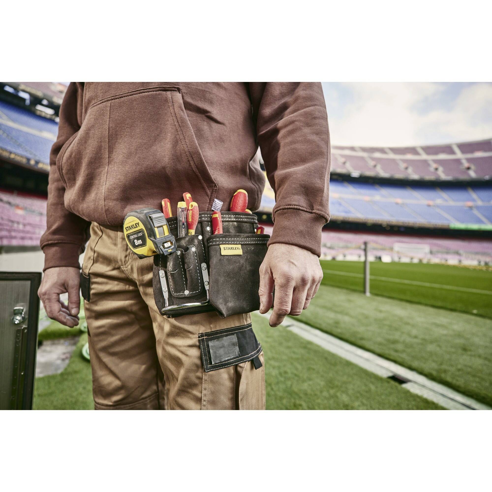 Osoba nosi torbę narzędziową z różnymi narzędziami przewieszoną na biodrze, stojąc na boisku piłkarskim przy linii bocznej.