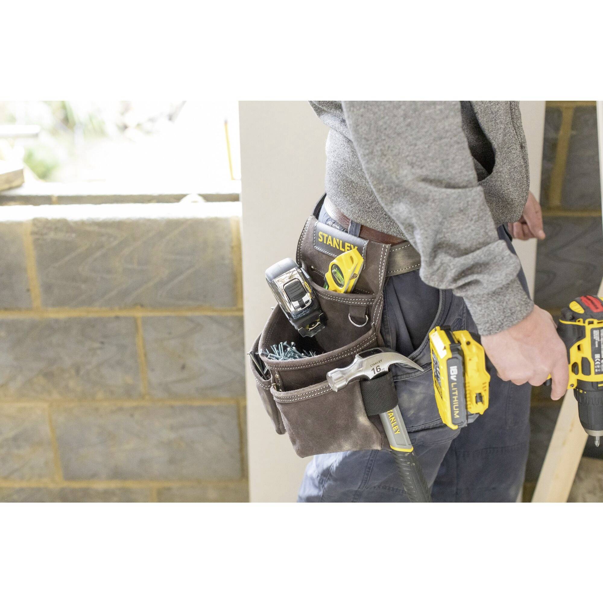 Osoba pracuje z wiertarką. Przy jej pasie wiszą młotek i różne narzędzia. W tle widać niedokończoną ścianę z cegły.