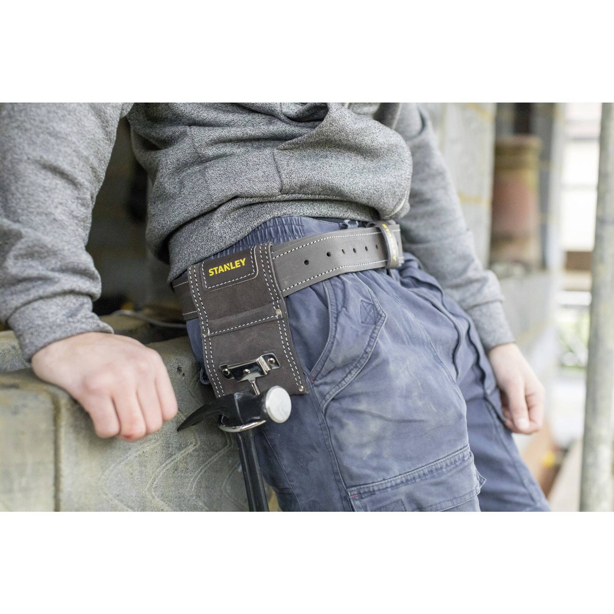 Osoba ubrana jest w niebieskie spodnie robocze i szary sweter, opierając się o ścianę. Do spodni przypięty jest pas na narzędzia.