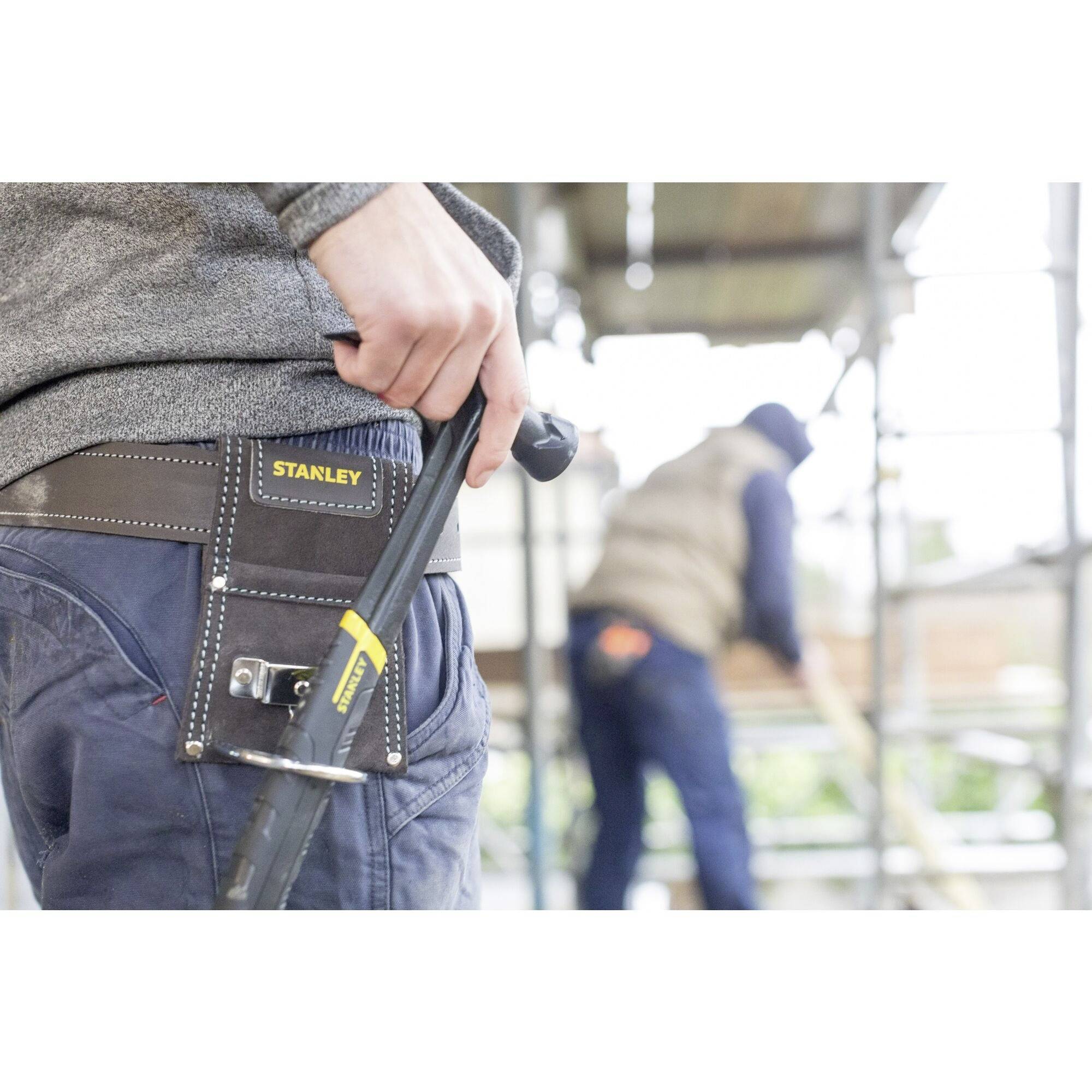 Robotnik budowlany stoi na pierwszym planie z narzędziami w torbie zawieszonej na pasie. W tle, nieostra sylwetka kolejnego pracownika wykonuje swoją pracę.