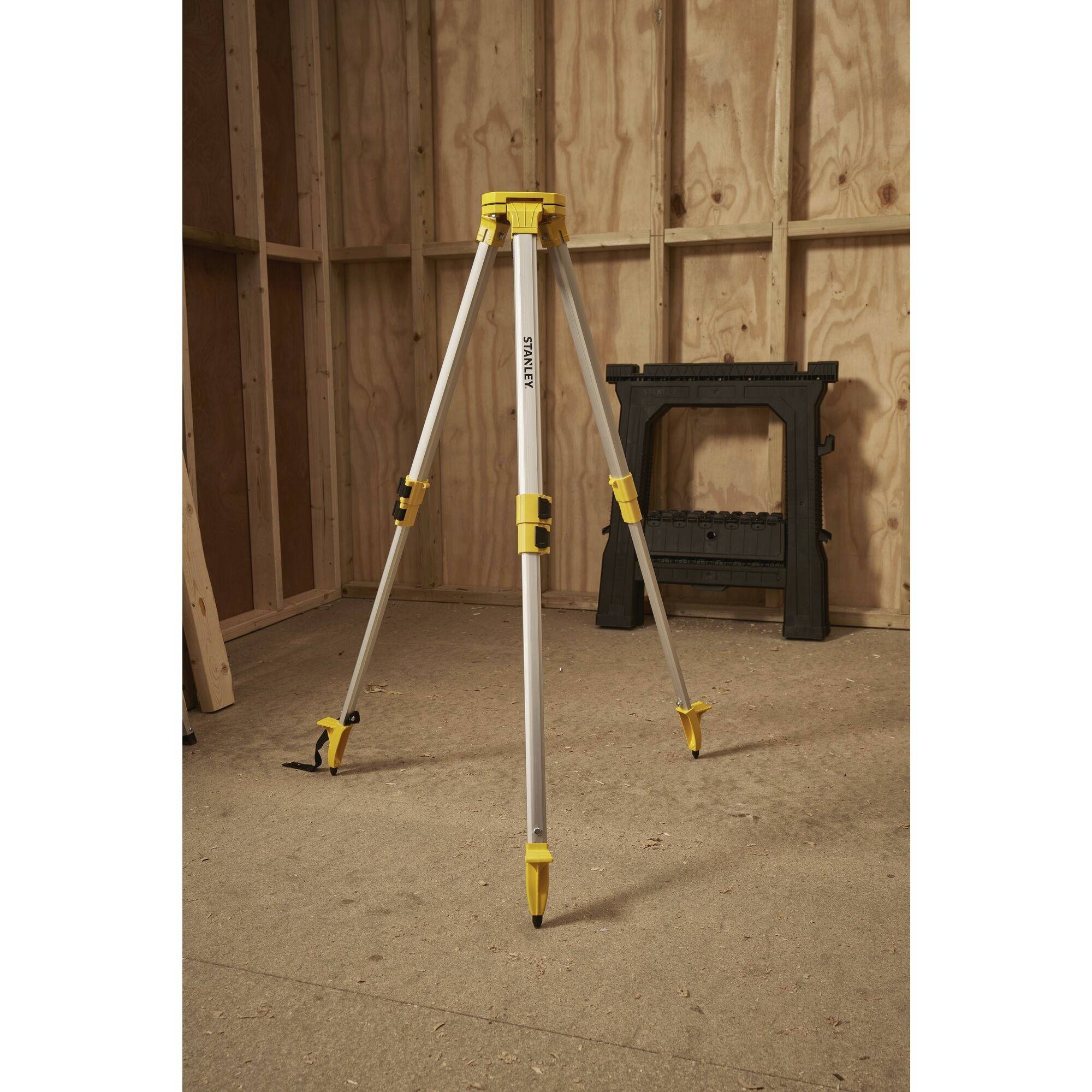 Srebrny, trójnożny statyw z żółtymi detalami stoi w niedokończonym drewnianym pomieszczeniu na drewnianej podłodze, obok czarnego wspornika.