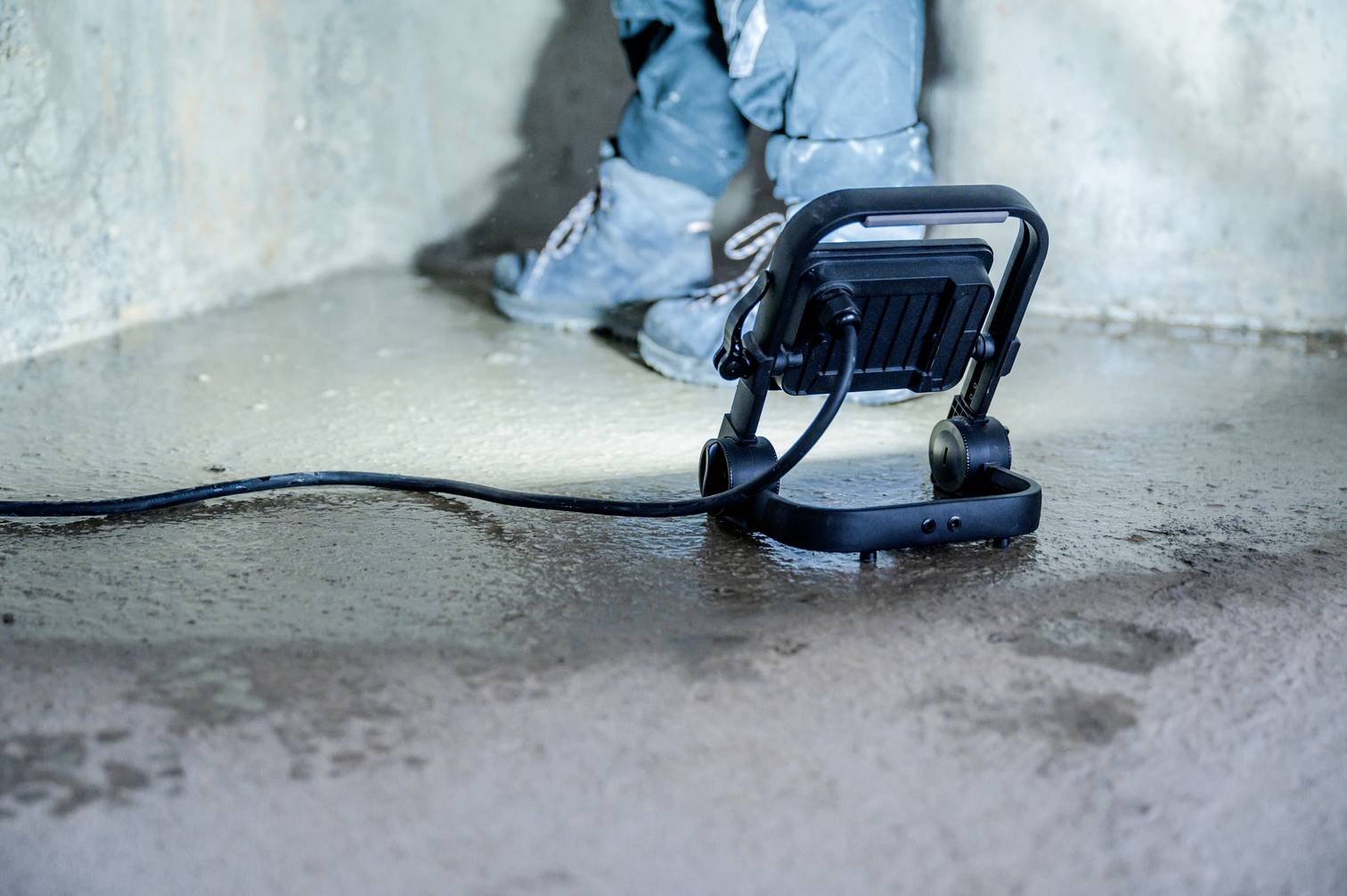 Przenośna lampa robocza oświetla betonową podłogę. W tle widoczne są nogi robotników w odzieży ochronnej.