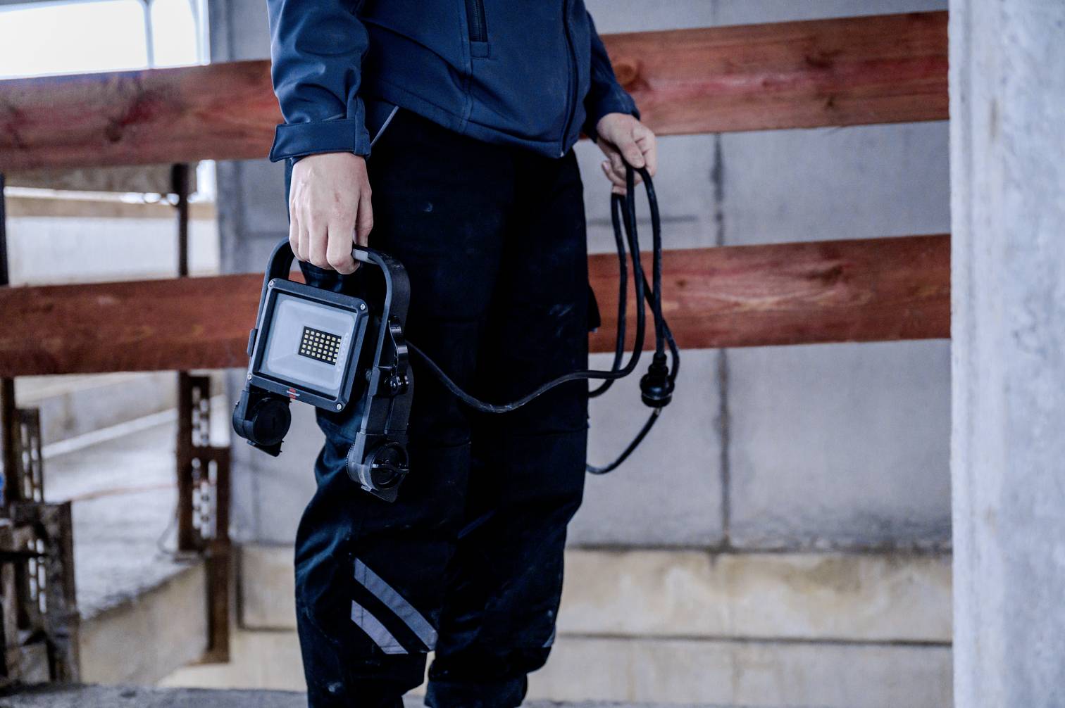 Pracownik w ubraniu roboczym trzyma w ręku przenośny reflektor budowlany, stojąc w niedokończonym budynku z betonowymi i drewnianymi elementami konstrukcyjnymi.