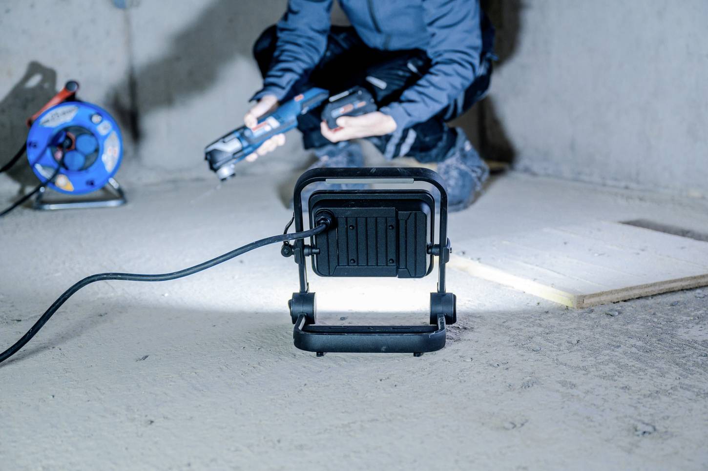 Osoba pracuje klęcząc na betonowej podłodze, posługując się narzędziem. Na pierwszym planie stoi włączony reflektor budowlany, oświetlający okolicę.