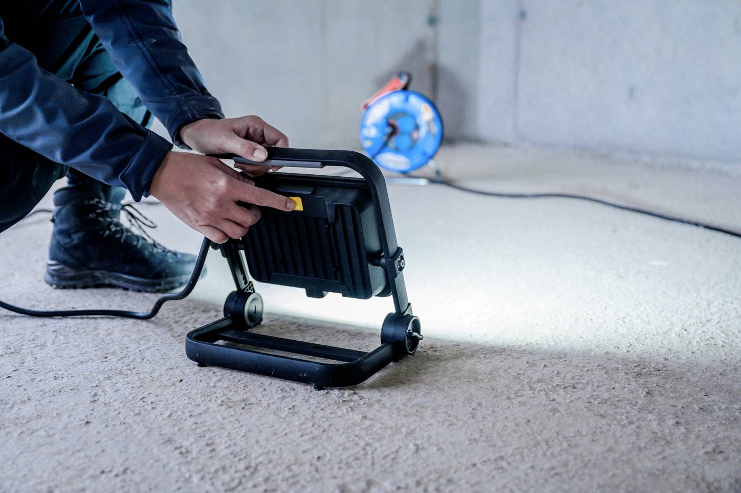 Osoba ustawia przenośny reflektor budowlany na betonowej podłodze. W tle widoczna jest rolka kabla.