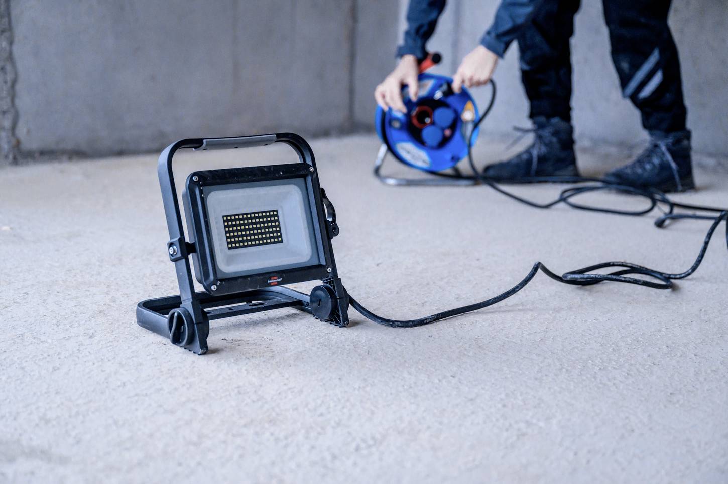 Reflektor budowlany stojący na podłodze jest połączony długim przewodem z bębnem kablowym. W tle widać osobę trzymającą tę bębnową rolkę.