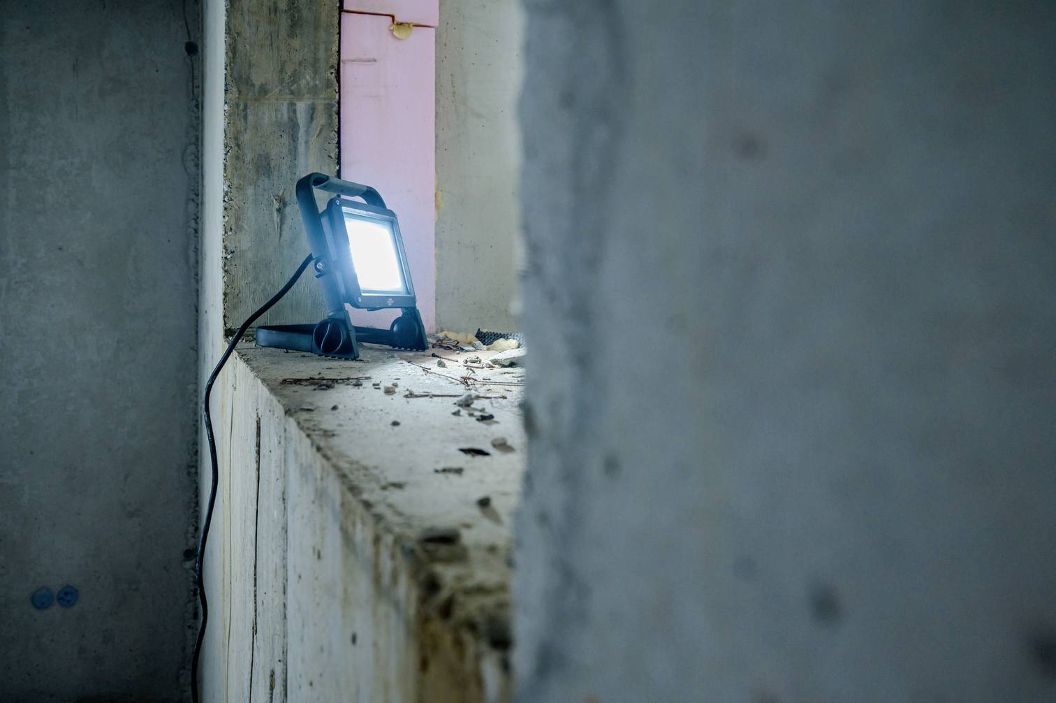 Reflektor budowlany oświetla zniszczone okienne skrzydło w opuszczonym pomieszczeniu. Widoczny jest kurz i gruz.