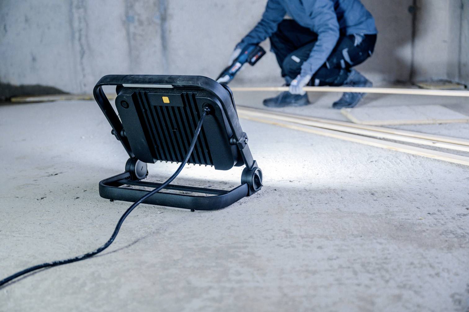 Osoba w ubraniu roboczym używa narzędzia w niedokończonym pomieszczeniu. Na pierwszym planie znajduje się lampa robocza oświetlająca okolicę.