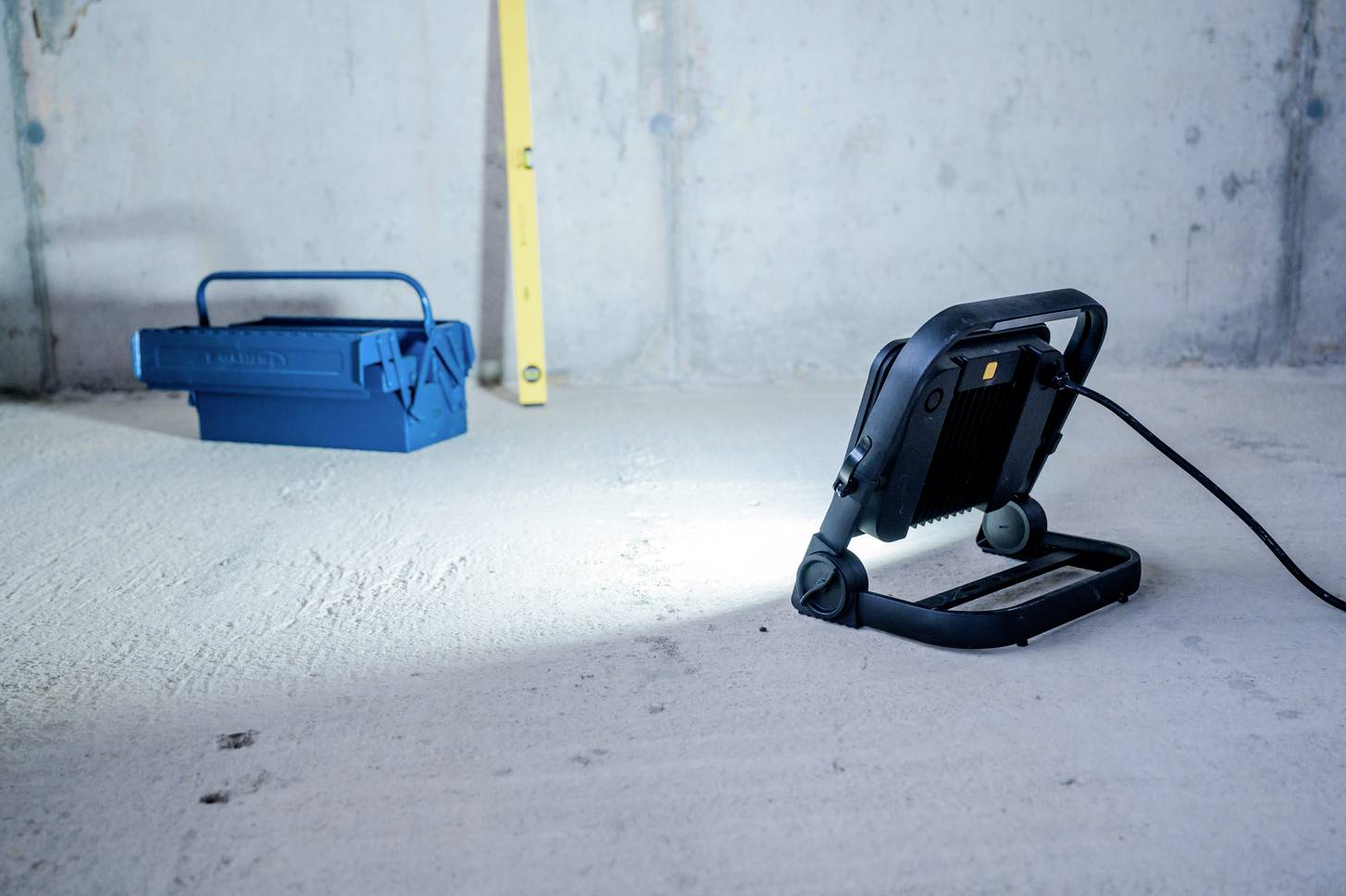 Niebieska skrzynka narzędziowa i lampa robocza na betonowej podłodze w niedokończonym pomieszczeniu. Lampa rzuca światło na podłogę.