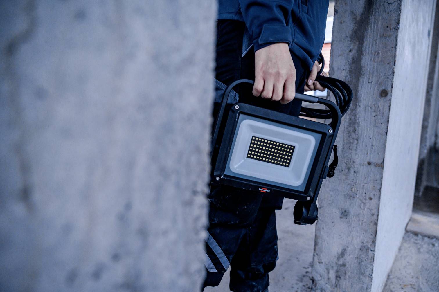 Osoba trzyma przenośny LED-owy reflektor budowlany. W tle widać betonowe ściany, co wskazuje na plac budowy.