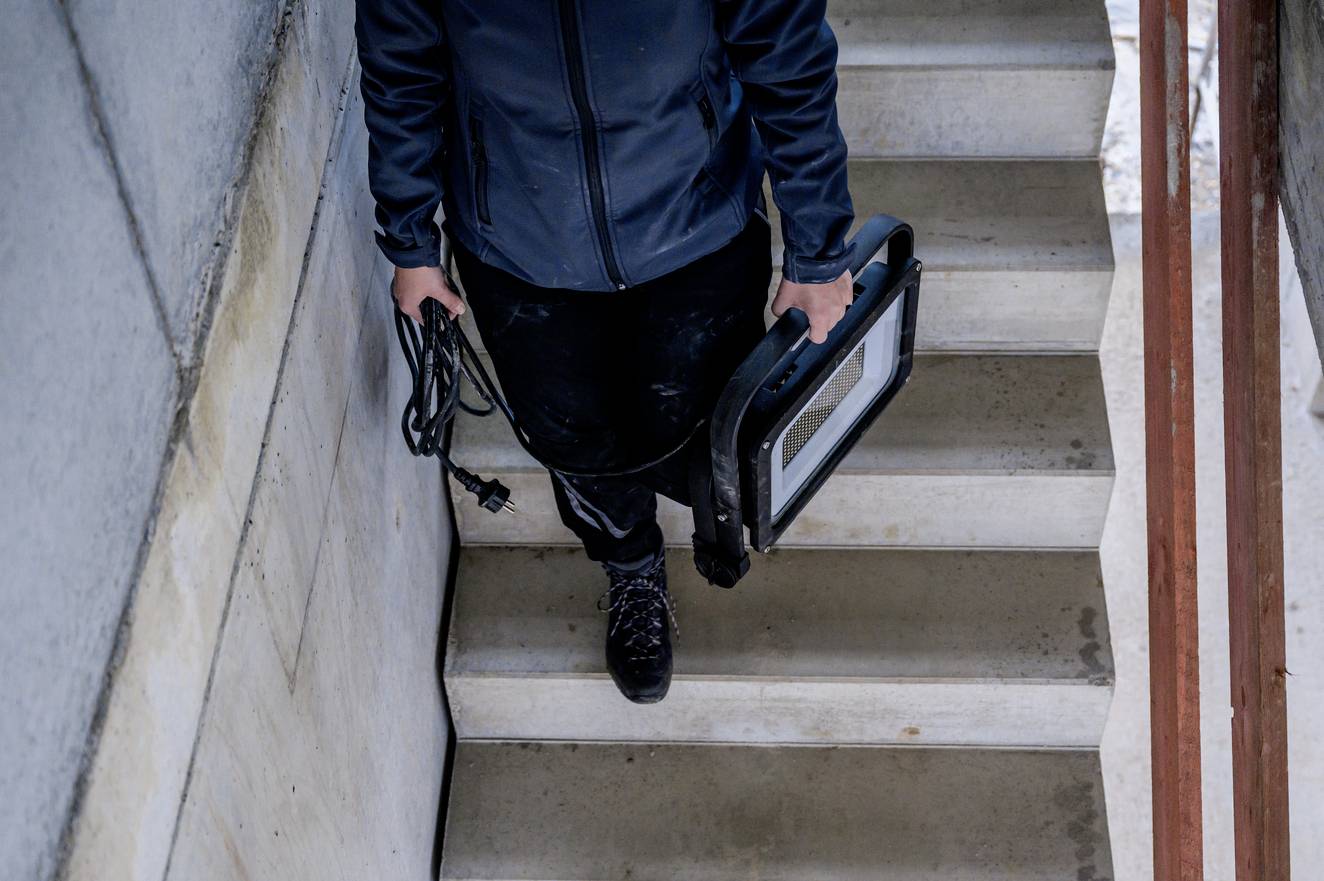 Osoba schodzi po schodach, trzymając w lewej ręce lampę roboczą, a w prawej przewód. Ubranie jest zimowe.