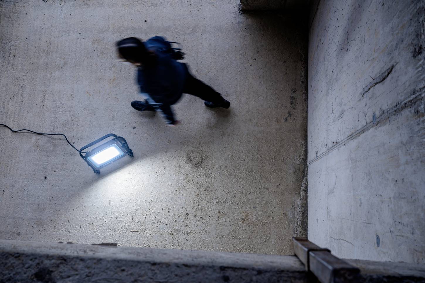 Osoba przechodzi obok włączonej lampy roboczej leżącej na betonowej nawierzchni.