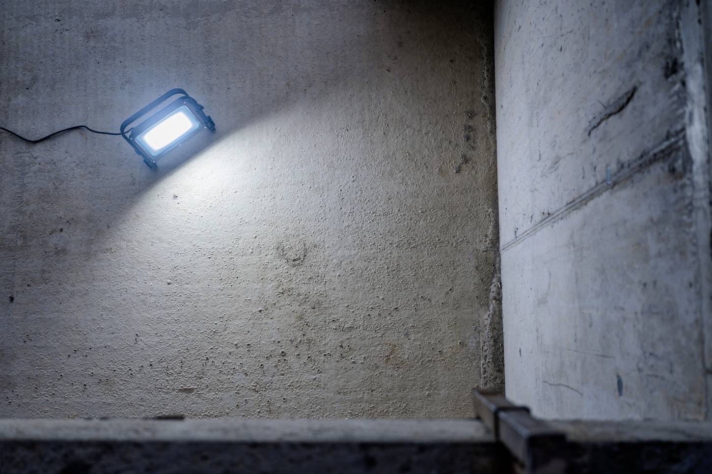 Niebieski reflektor budowlany oświetla szarą betonową ścianę w pustym pomieszczeniu. Kabel biegnie w lewą stronę.