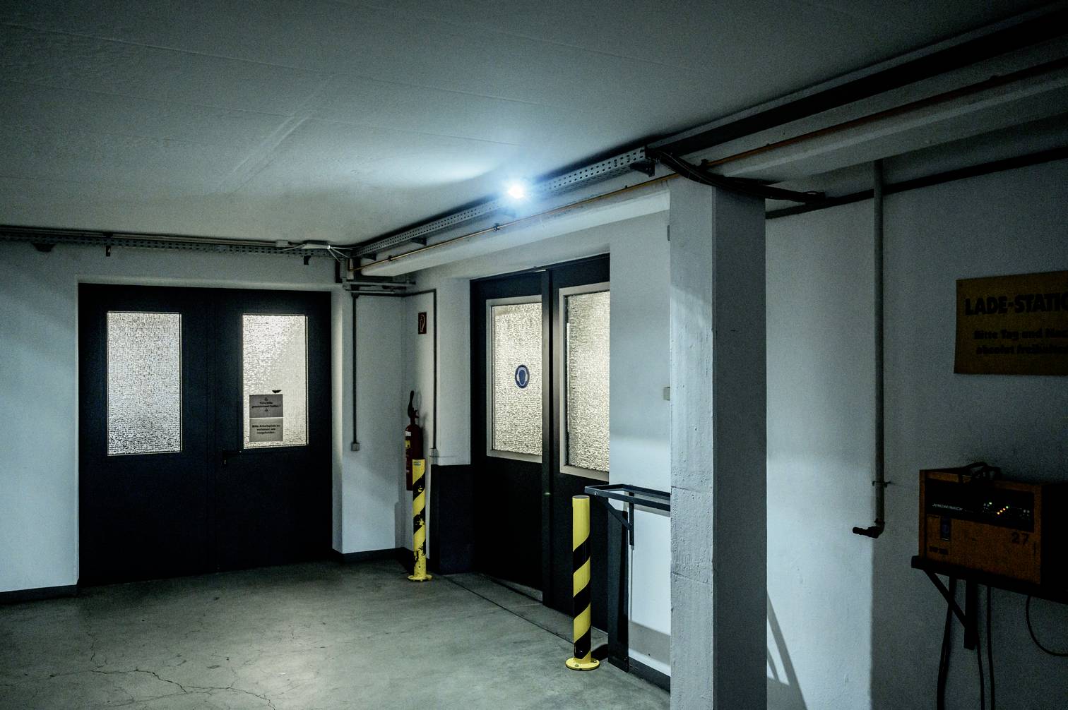 Parking wielopoziomowy z zamkniętymi drzwiami, oświetlony niebieskim światłem. Obok stacji ładowania wisi tabliczka z napisem "Stacja ładowania".