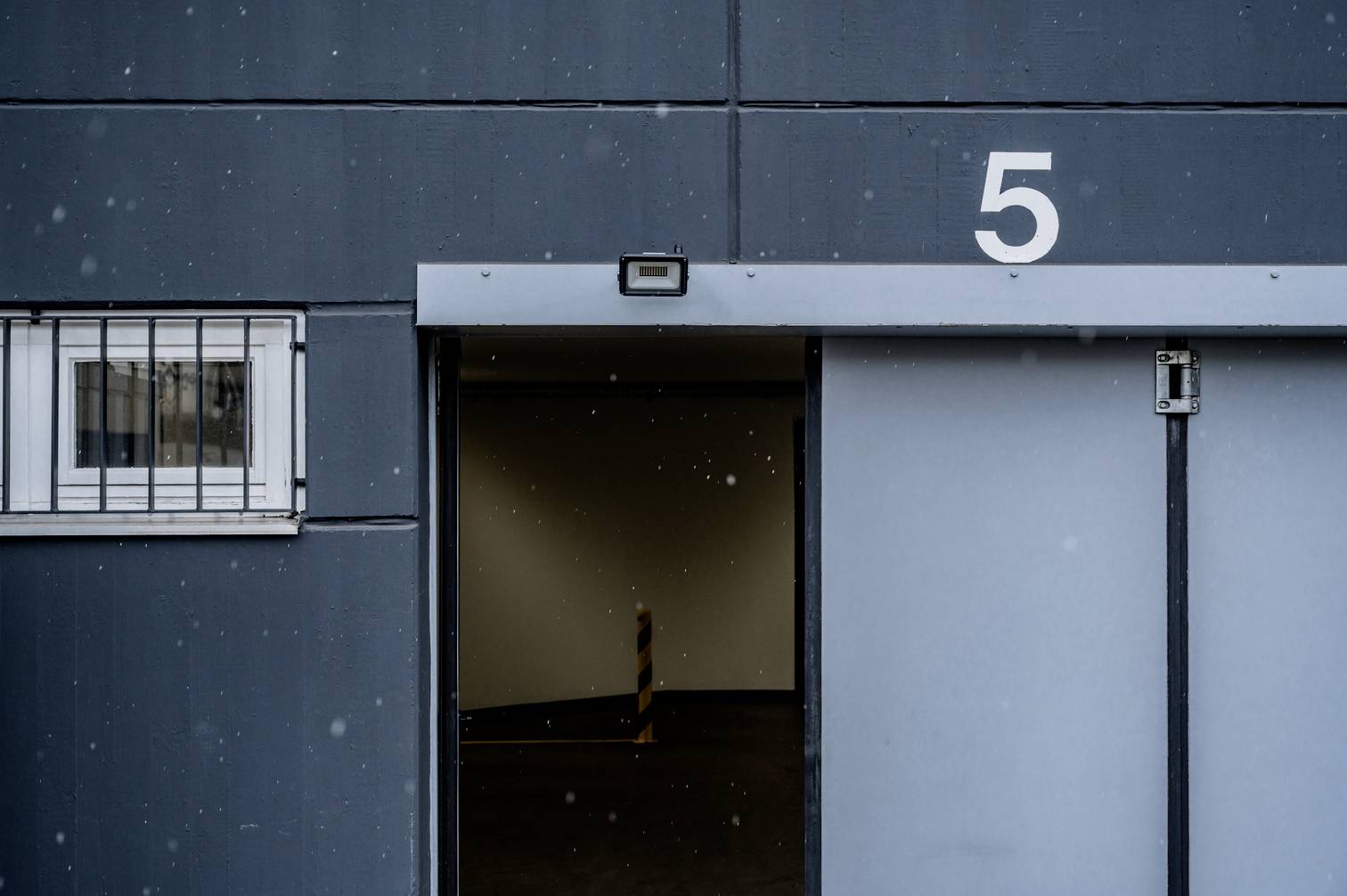 Zamknięte szare drzwi przesuwne z numerem 5, obok małe okno z kratą. Mimo padającego śniegu obraz tchn spokojnym nastrojem.