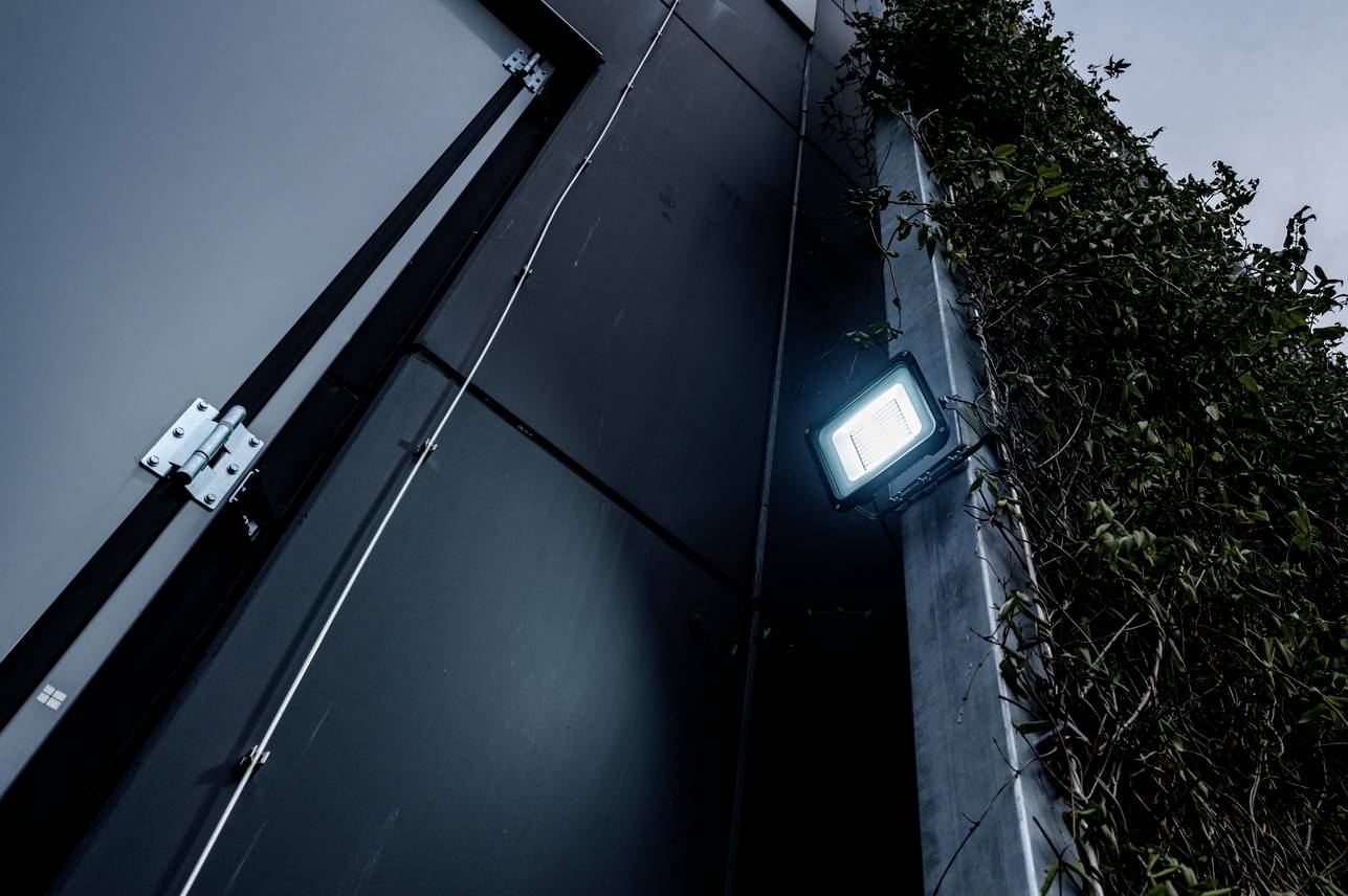 Widok zewnętrzny budynku o zmierzchu, jasno świecący reflektor na ścianie, po prawej stronie rośliny pną się w górę po budynku.