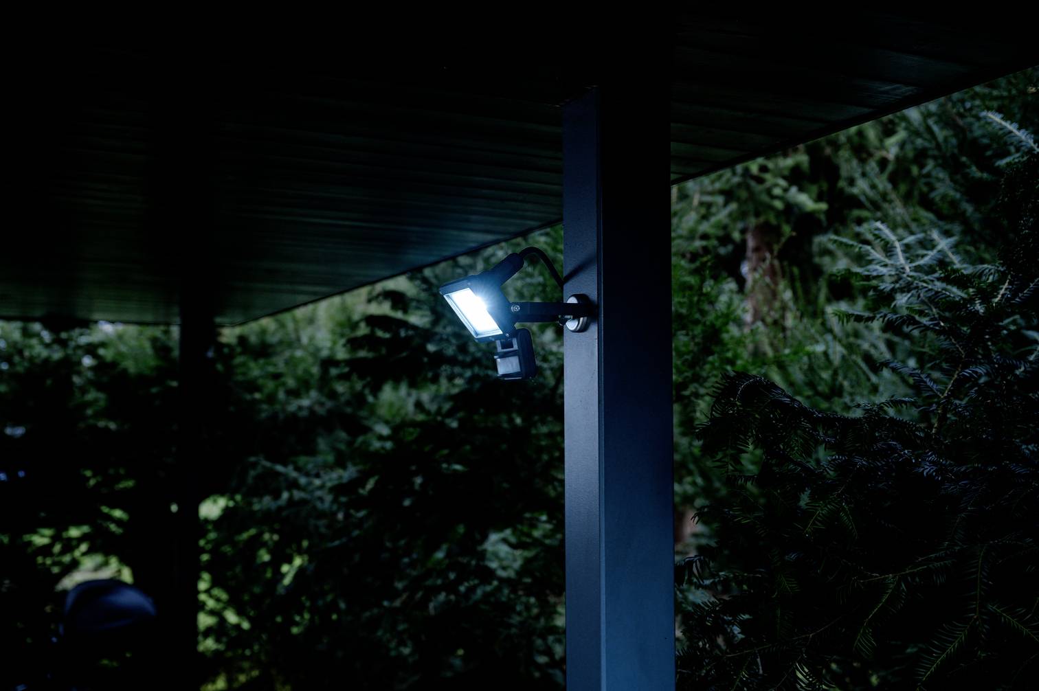 Lampa zewnętrzna oświetla nocą obszar ogrodu, otoczony drzewami.