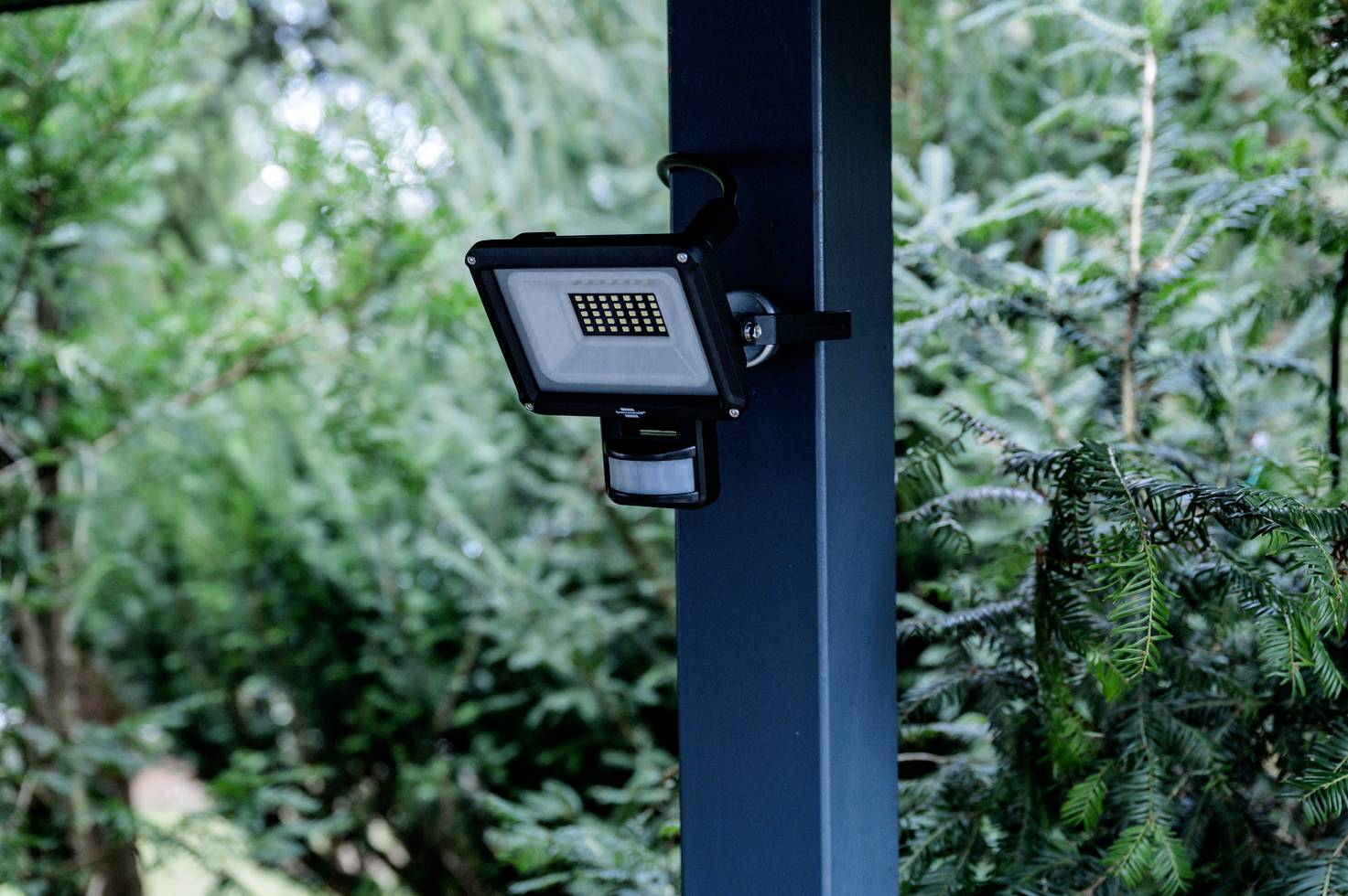 Czujnik ruchu z lampką bezpieczeństwa zamontowany na słupie, otoczony zielenią liści, przeznaczony do monitoringu terenu zewnętrznego.