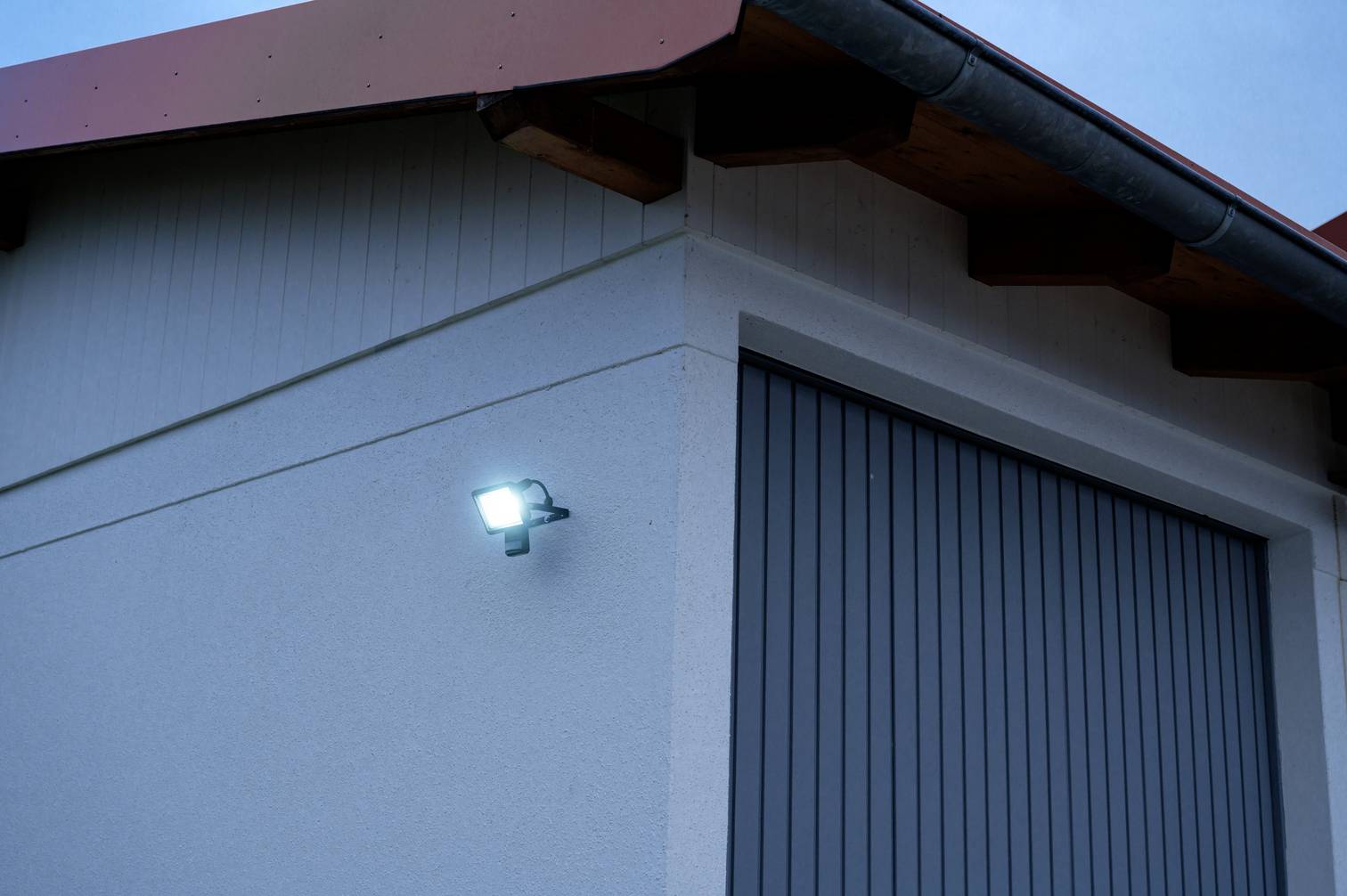 Włączone światło zewnętrzne budynku oświetla białą ścianę i szarą bramę garażową o zmierzchu.
