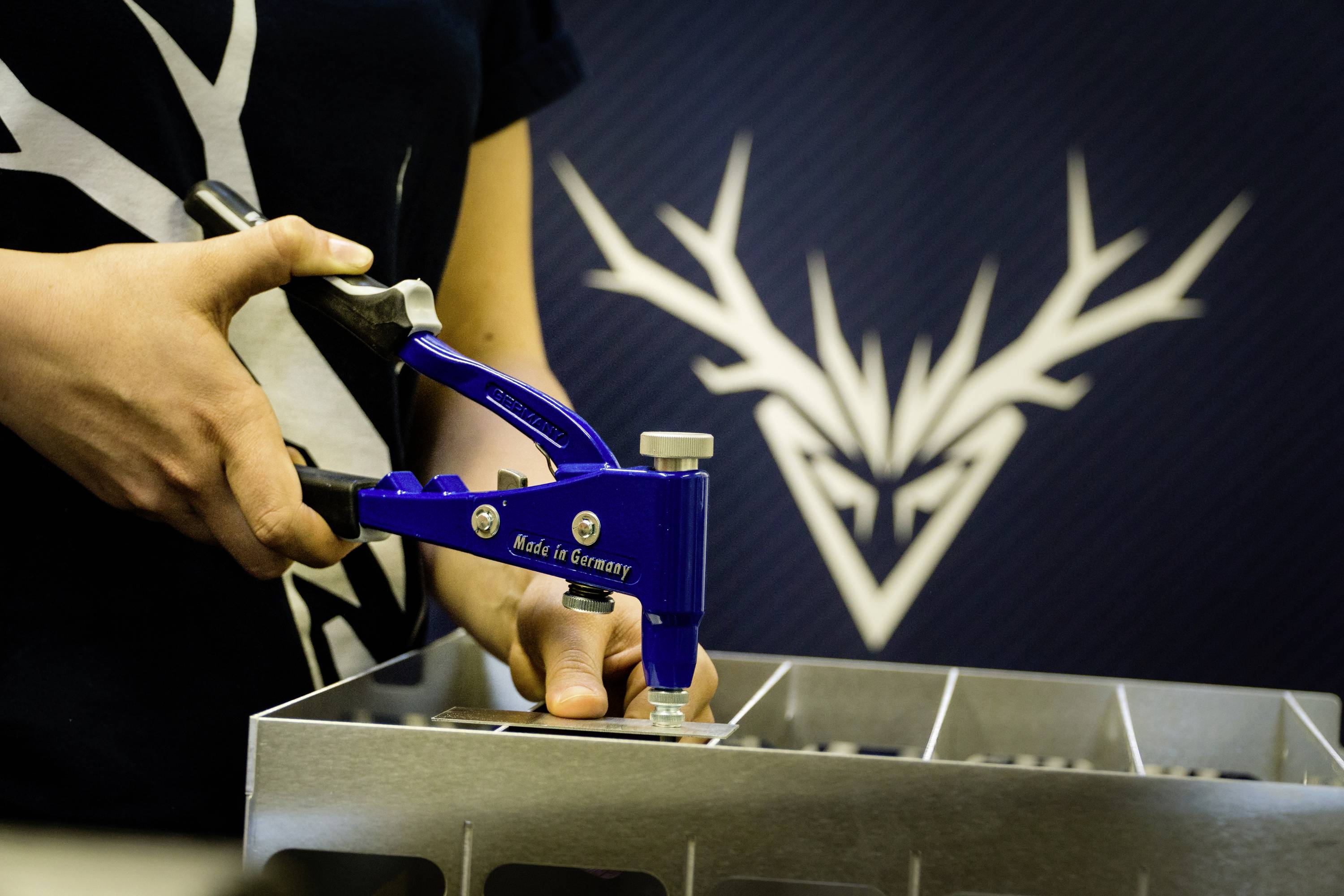 Osoba używa niebieskiego narzędzia do zamocowania czegoś. W tle widoczne jest logo z poroża jelenia.