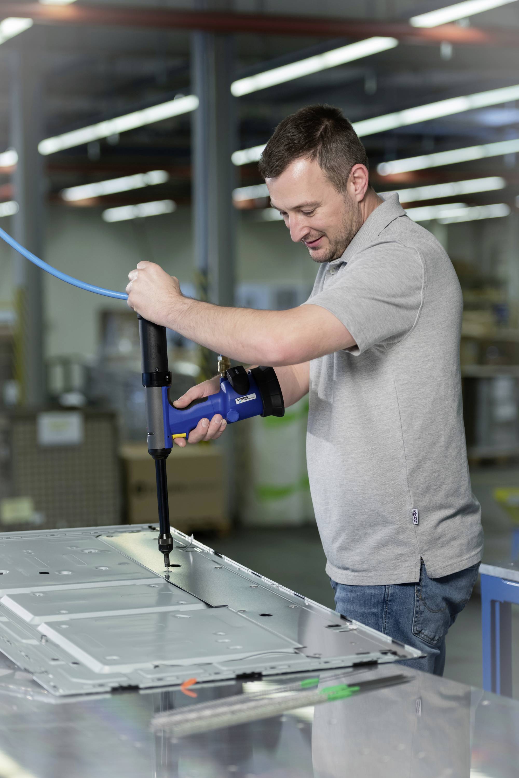 Mężczyzna w szarej koszulce polo mocuje blachę metalową za pomocą wiertarki w środowisku fabrycznym.