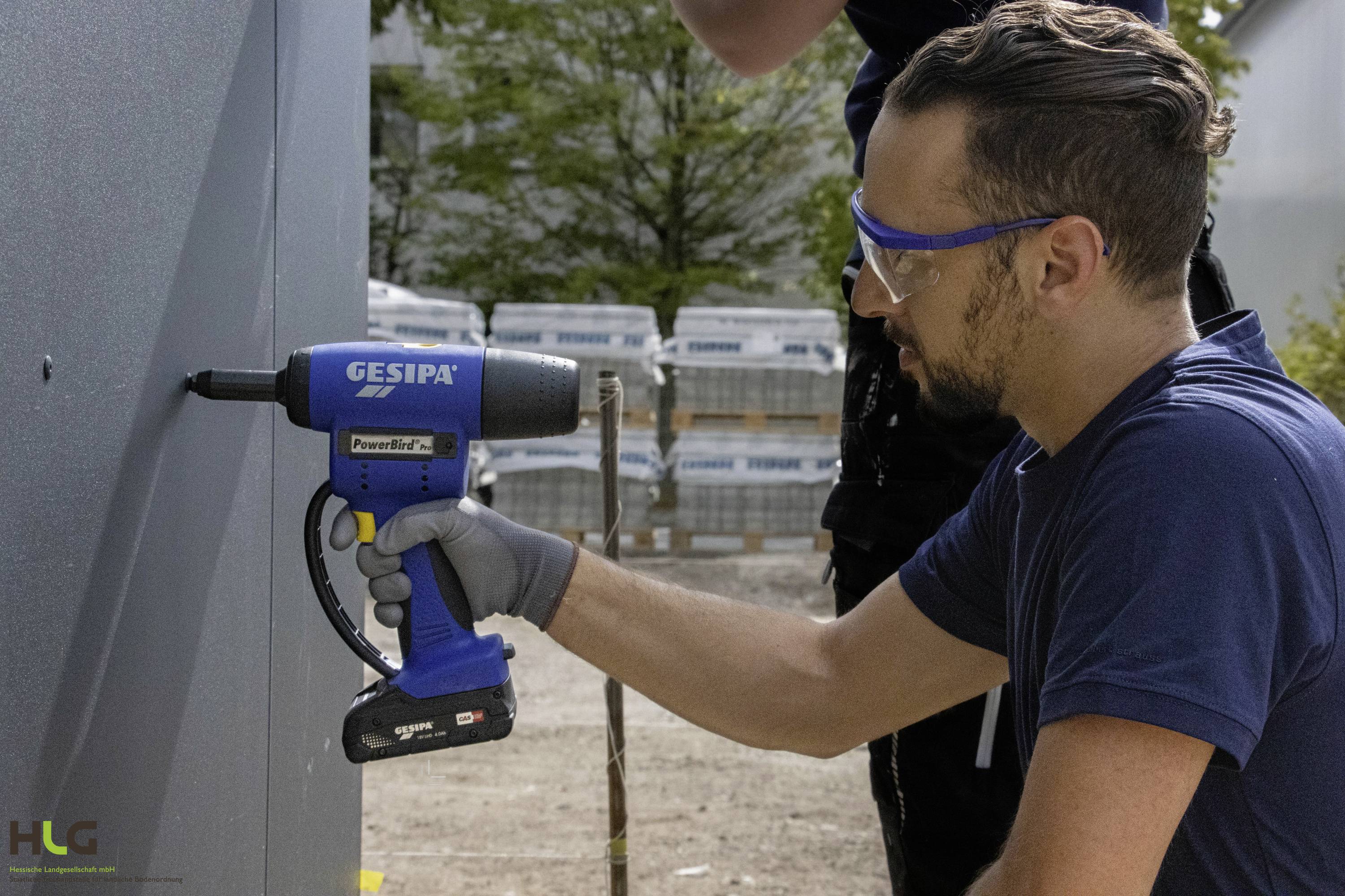 Mężczyzna montuje element na ścianie za pomocą niebieskiego elektronarzędzia. Nosi okulary ochronne i rękawice robocze.