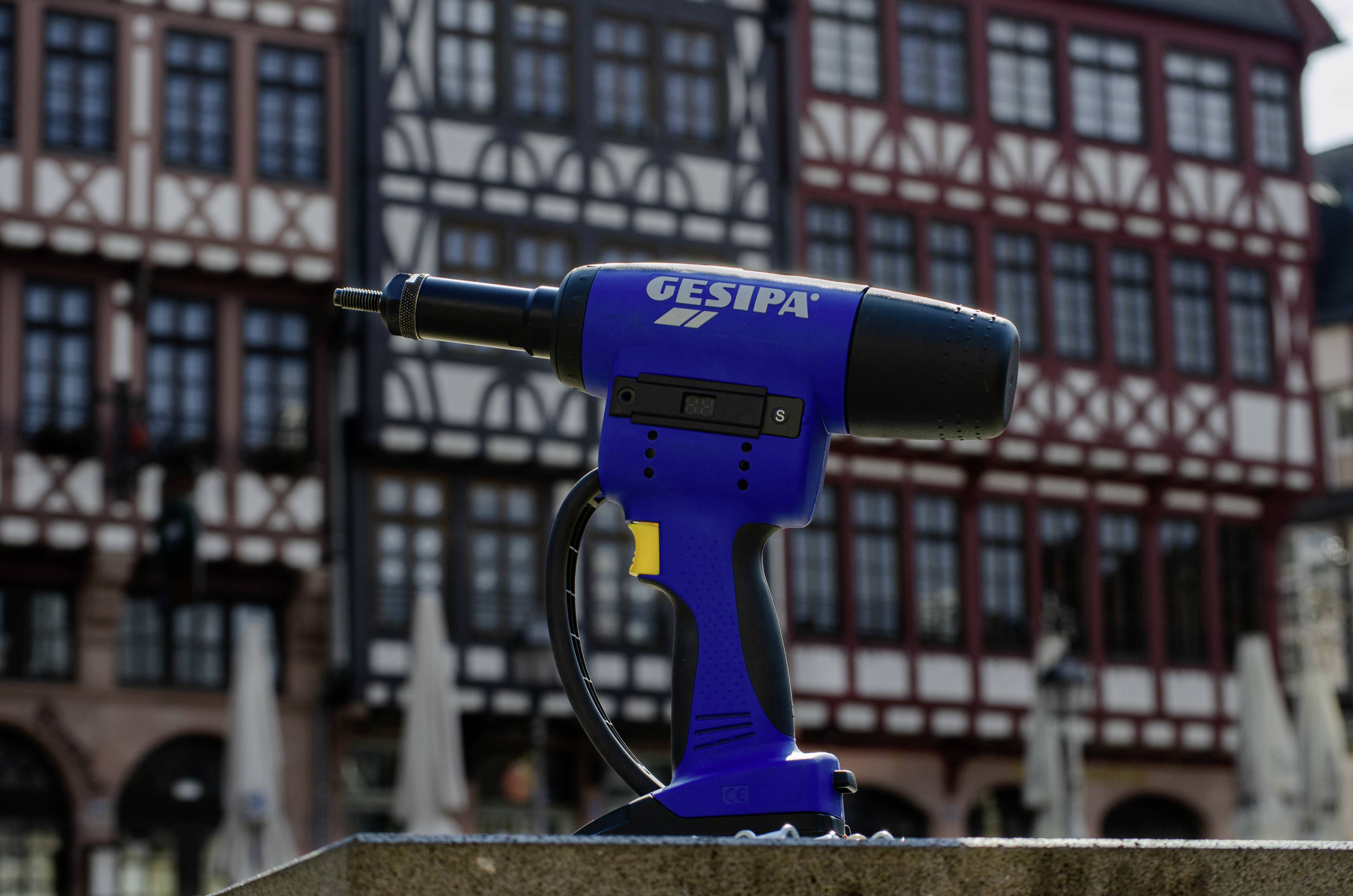 Niebieskie narzędzie z napisem 'Gesipa' na pierwszym planie. W tle widoczne są domy w stylu szachulcowym.