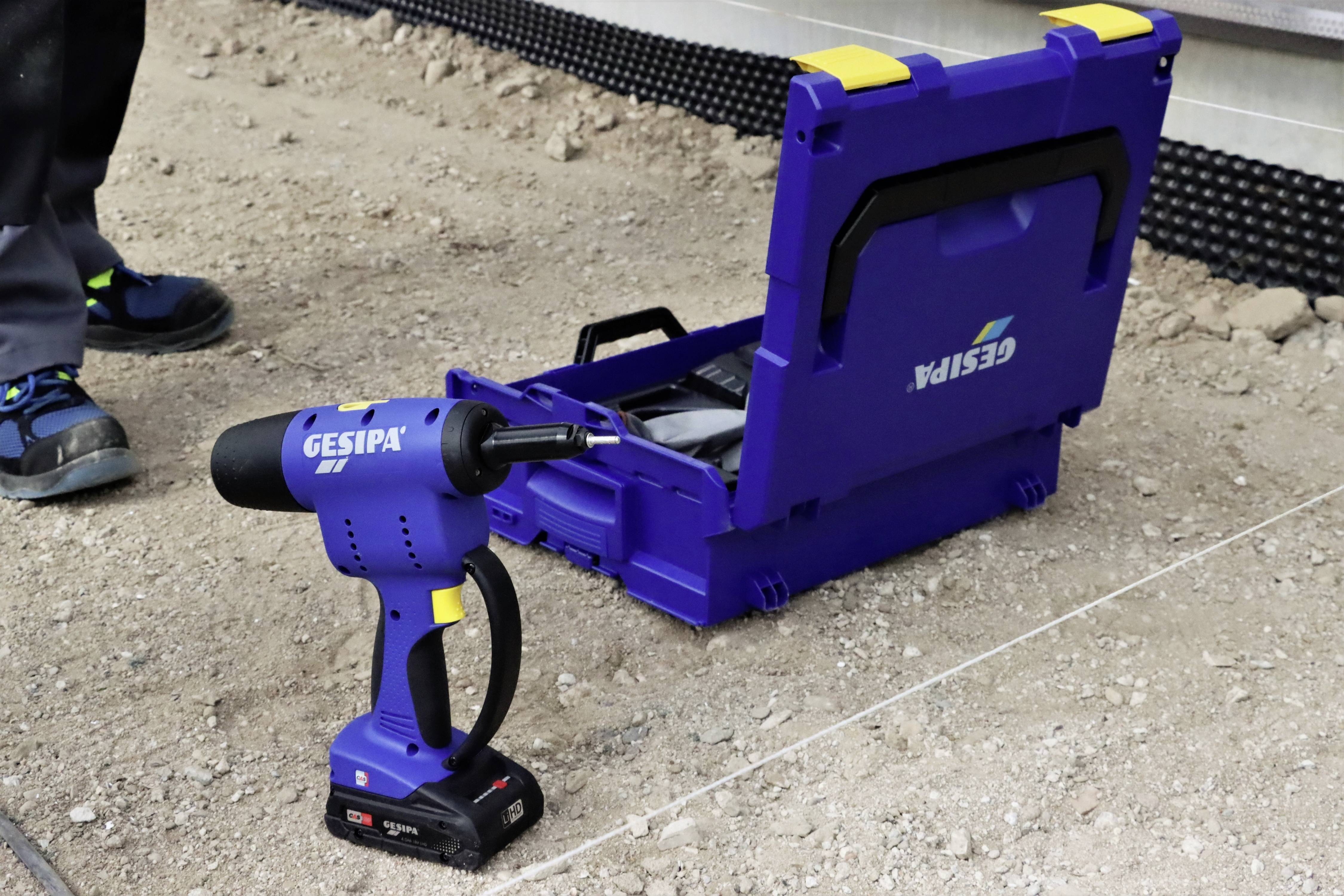 Niebieski akumulatorowy riveter marki Gesipa stoi na podłodze budowy obok otwartej niebieskiej walizki transportowej.