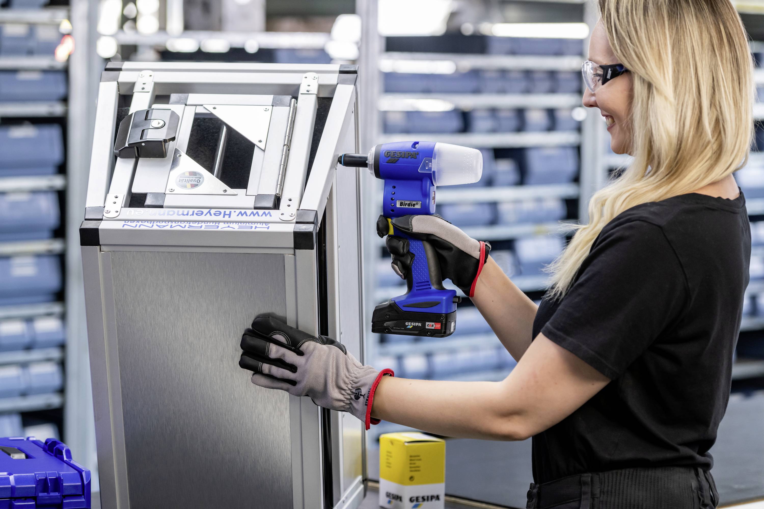 Kobieta w okularach ochronnych i rękawiczkach używa elektronarzędzia, aby zamknąć metalowe pudełko. W tle widoczne są regały z pudłami.